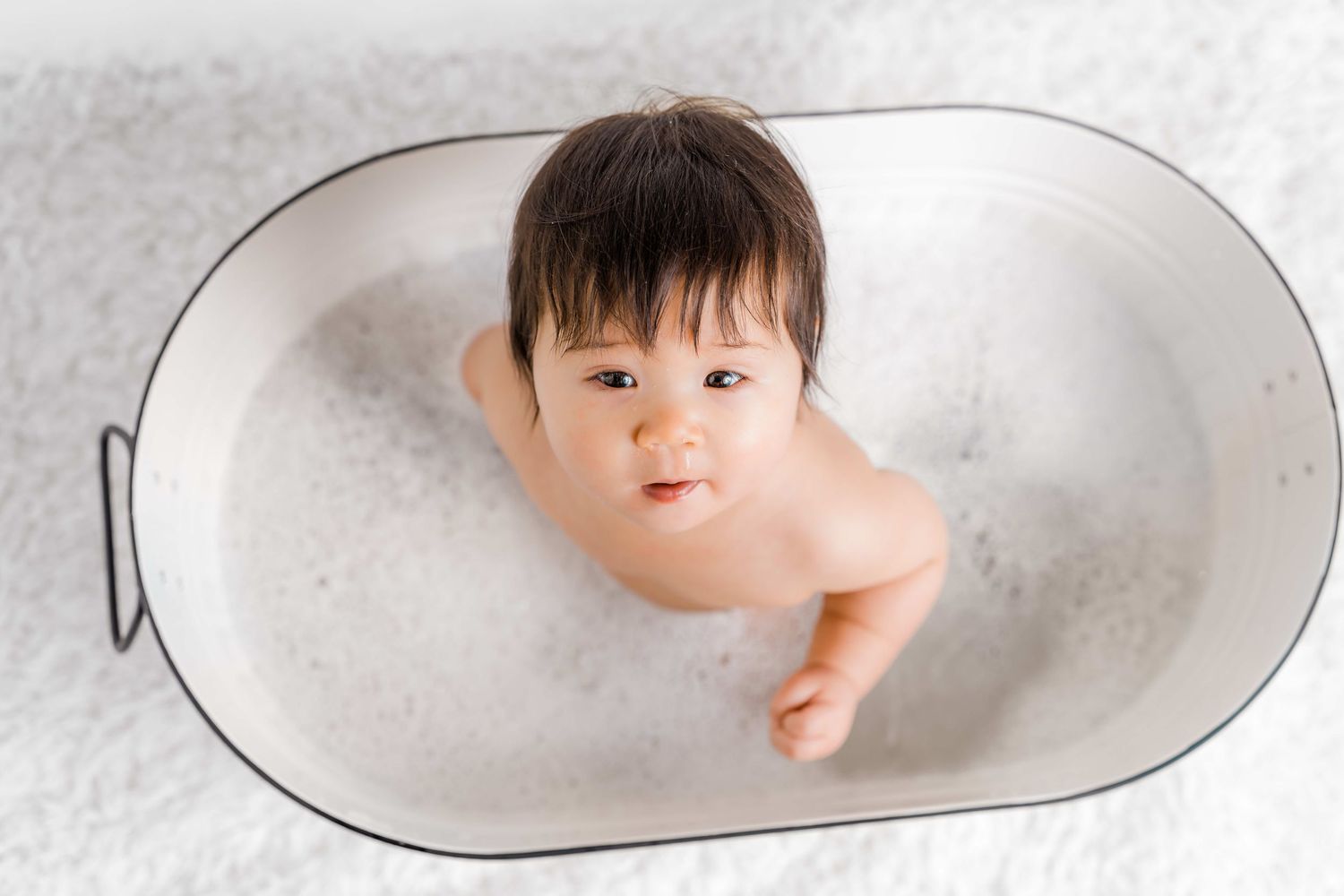 A baby takes a bubble bath in a white oval bathtub filled with soapy foam.