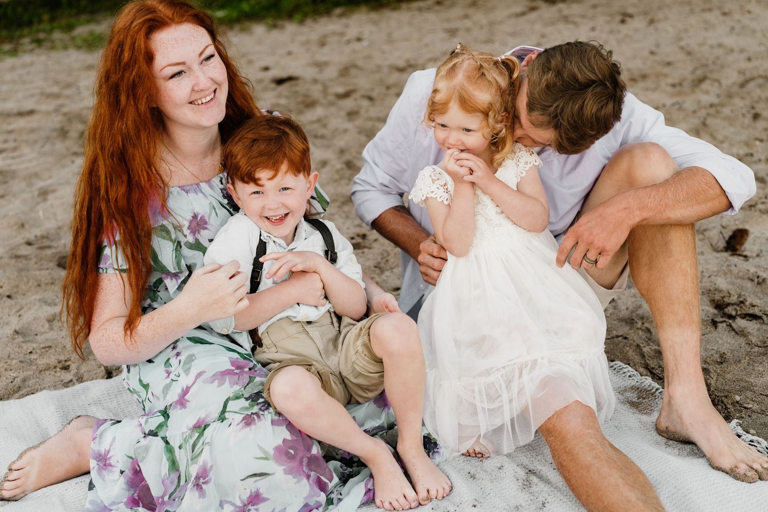 A happy family of four sits together on a sandy beach, smiling and laughing in elegant casual attire.