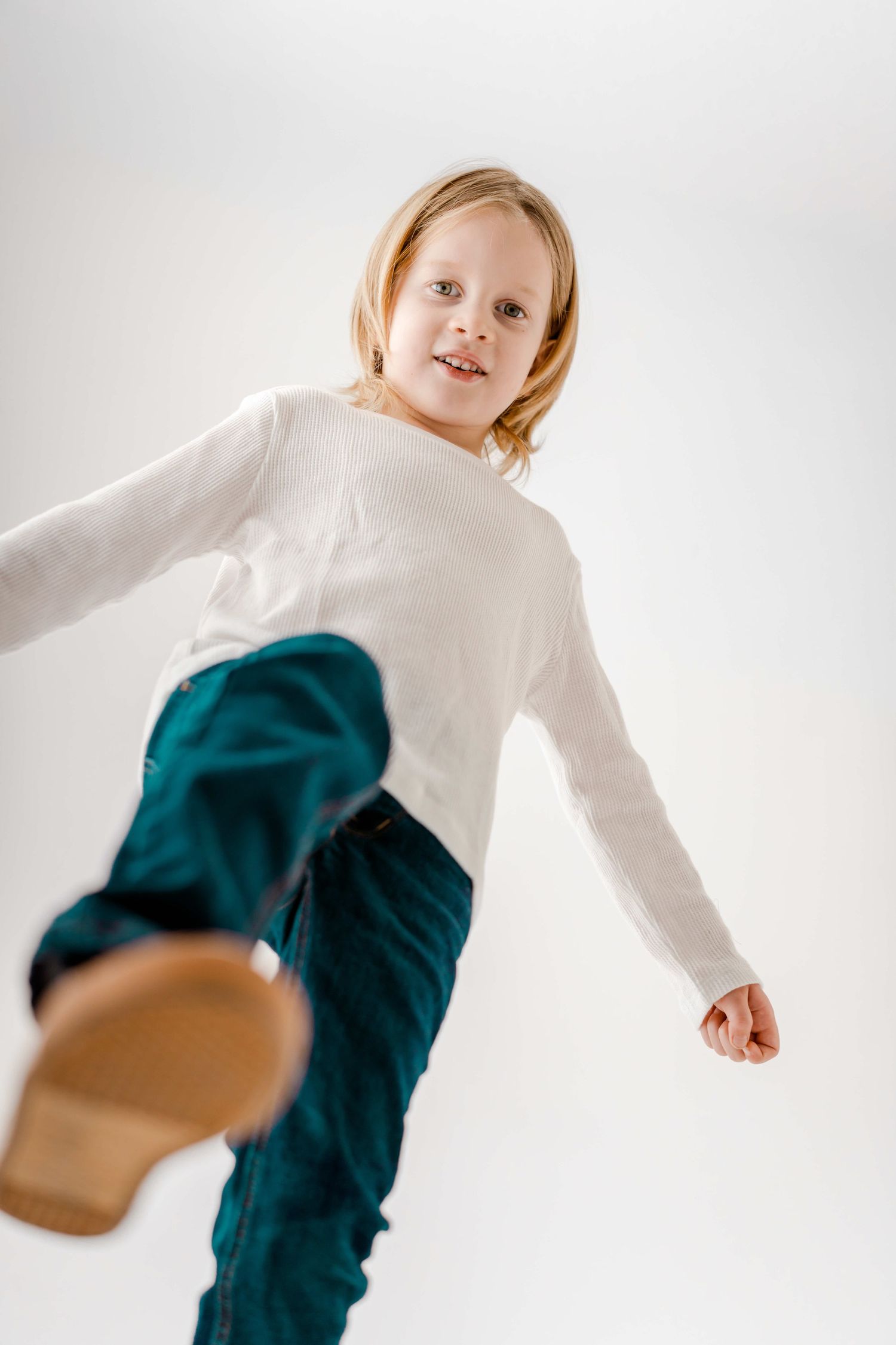 Child in white top and teal pants dances energetically in front of bright white studio background.