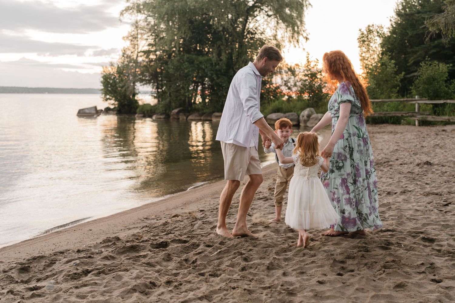 Family of four enjoying a sunset beach walk on sandy shore near calm lake waters surrounded by lush green trees.