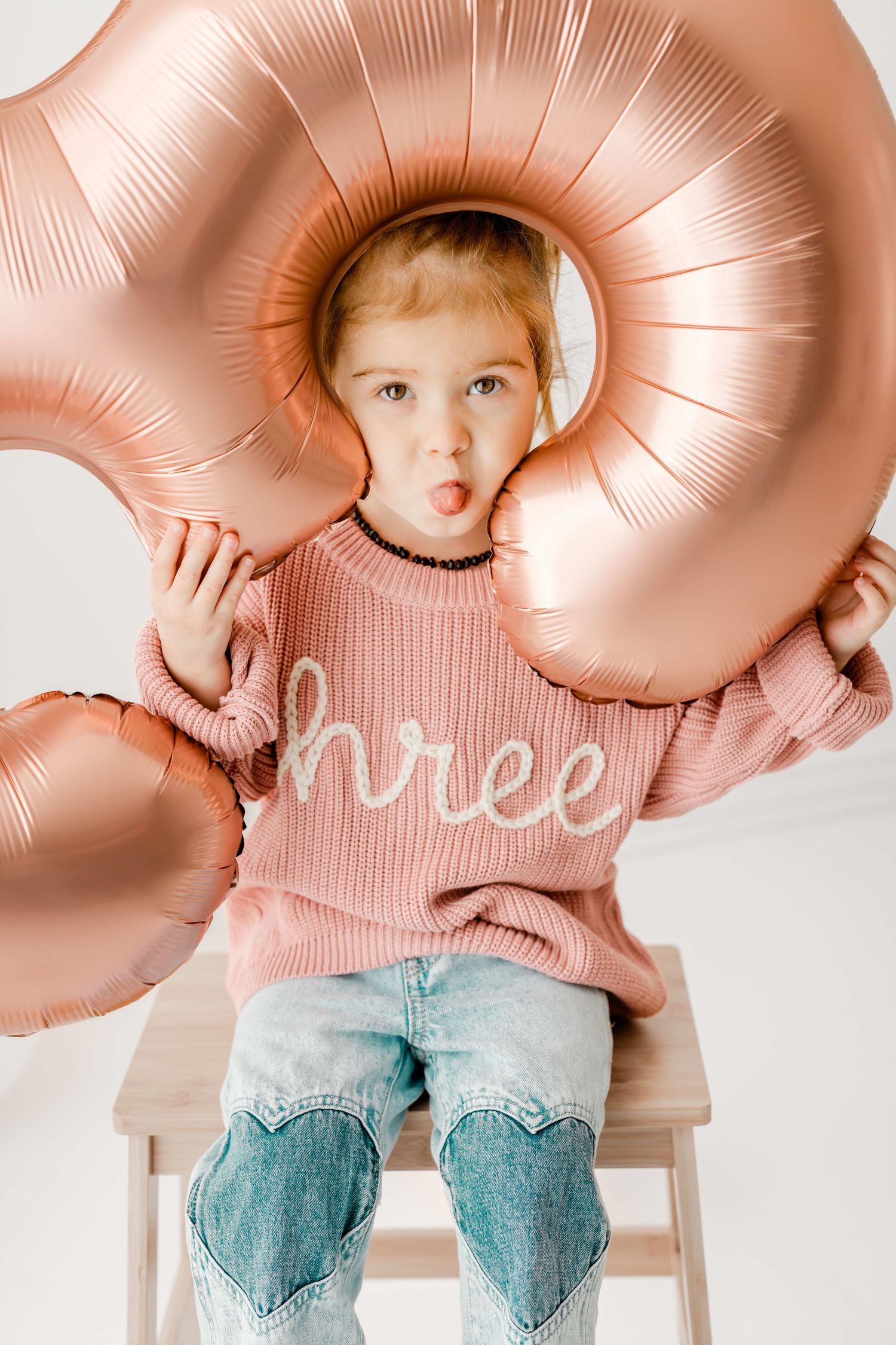 A child poses playfully through a rose gold number 3 balloon while wearing a pink sweater and heart-patched jeans.
