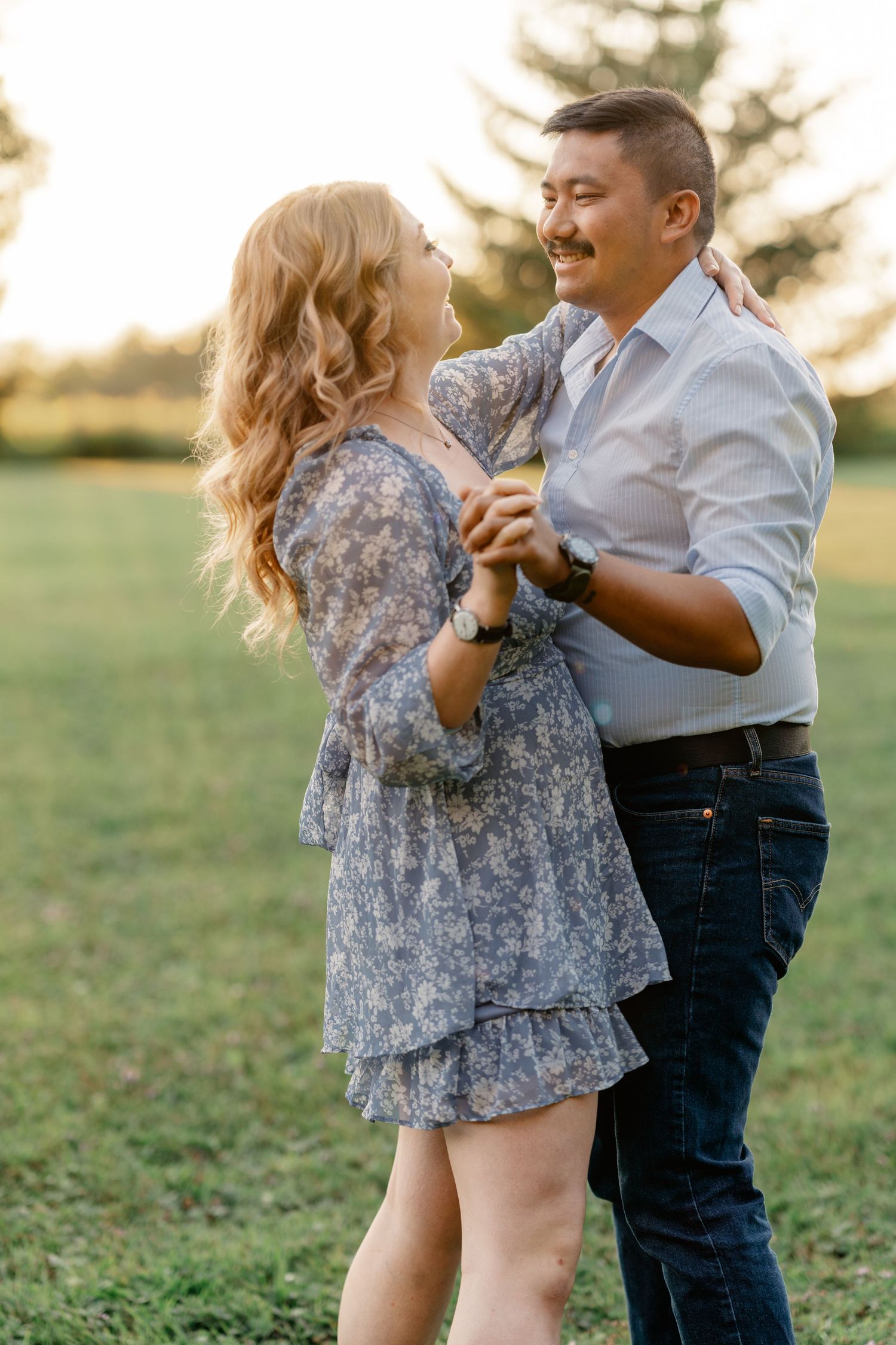 A couple dances together in a grassy field at sunset, the woman wearing a flowy floral dress and the man in jeans and white shirt.