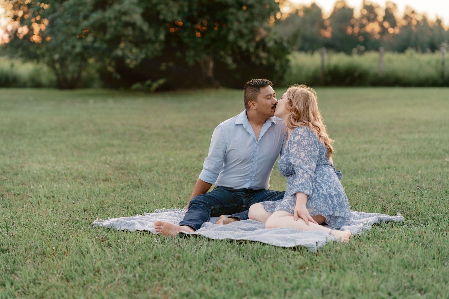 A couple shares a romantic moment while laying on a blanket in a grassy field at sunset.