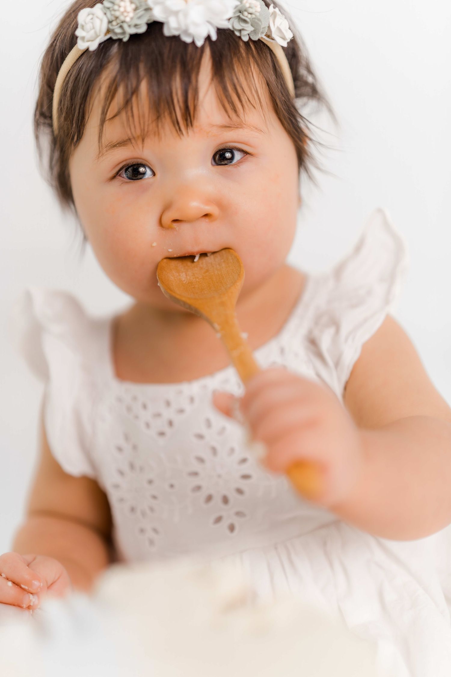 Sweet baby in white lace dress holds and chews on wooden teething toy against light background.
