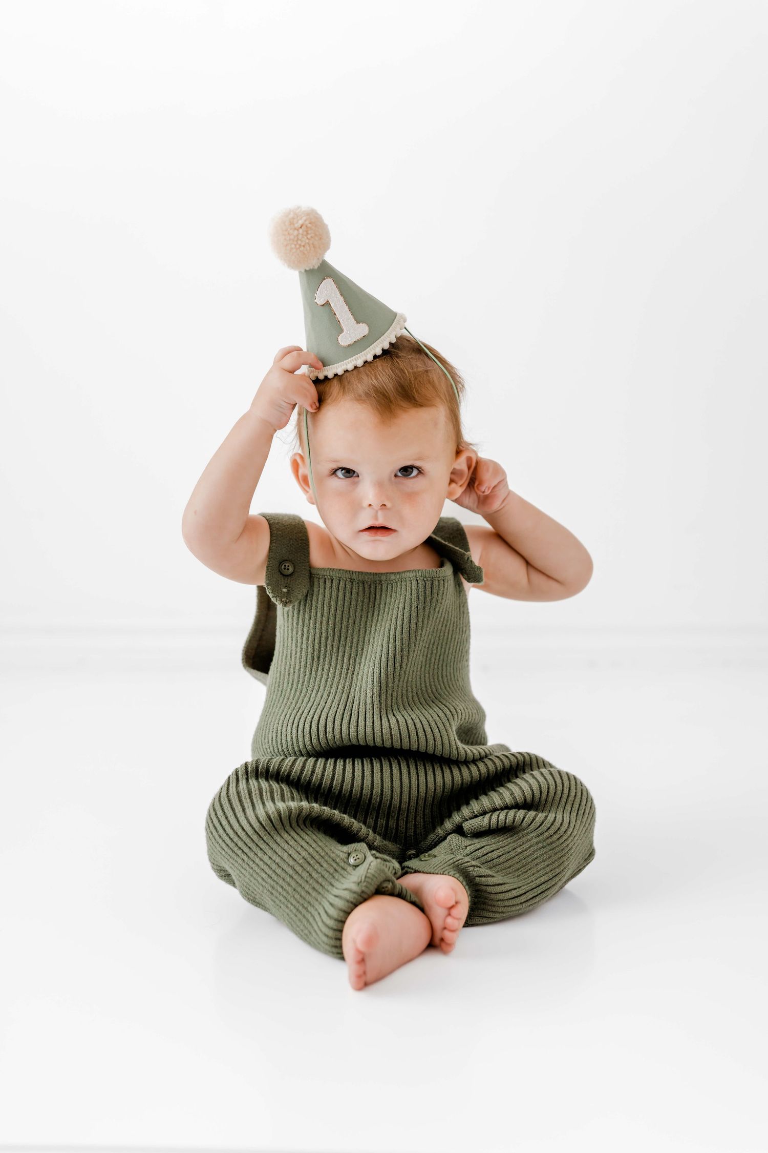Small child wearing olive green ribbed romper and party hat sits on white background in multiple poses.