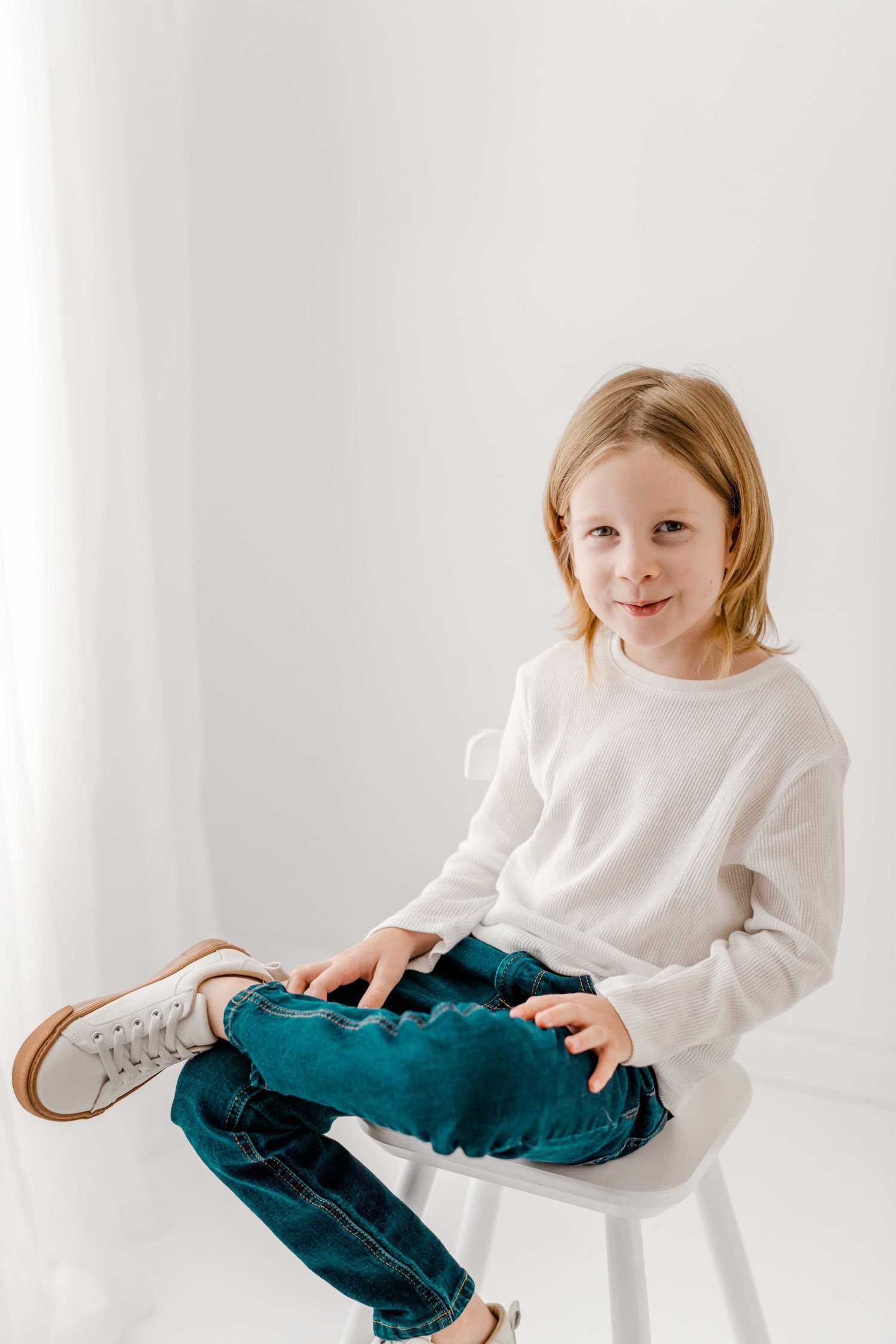 Child wearing white sweater and teal pants sits casually on white stool in minimalist setting.