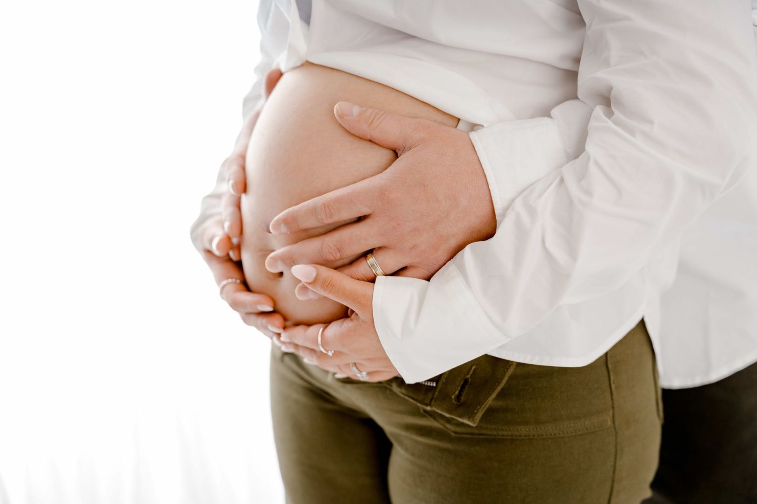 Pregnant woman in white shirt and olive pants cradles her baby bump while standing against a neutral background.