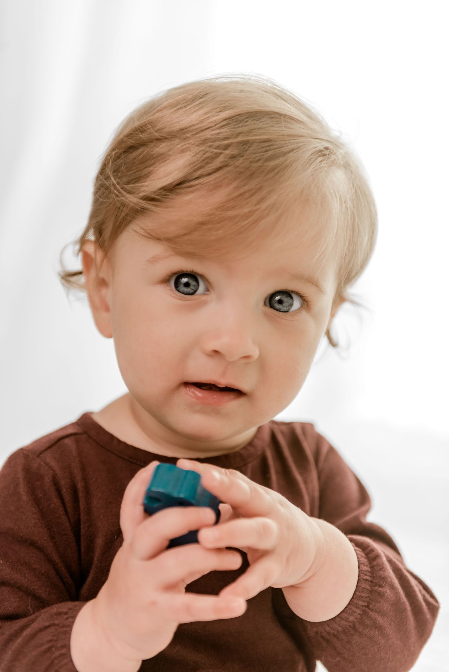 A young child in a brown shirt holds a small blue toy while looking up with wide eyes.