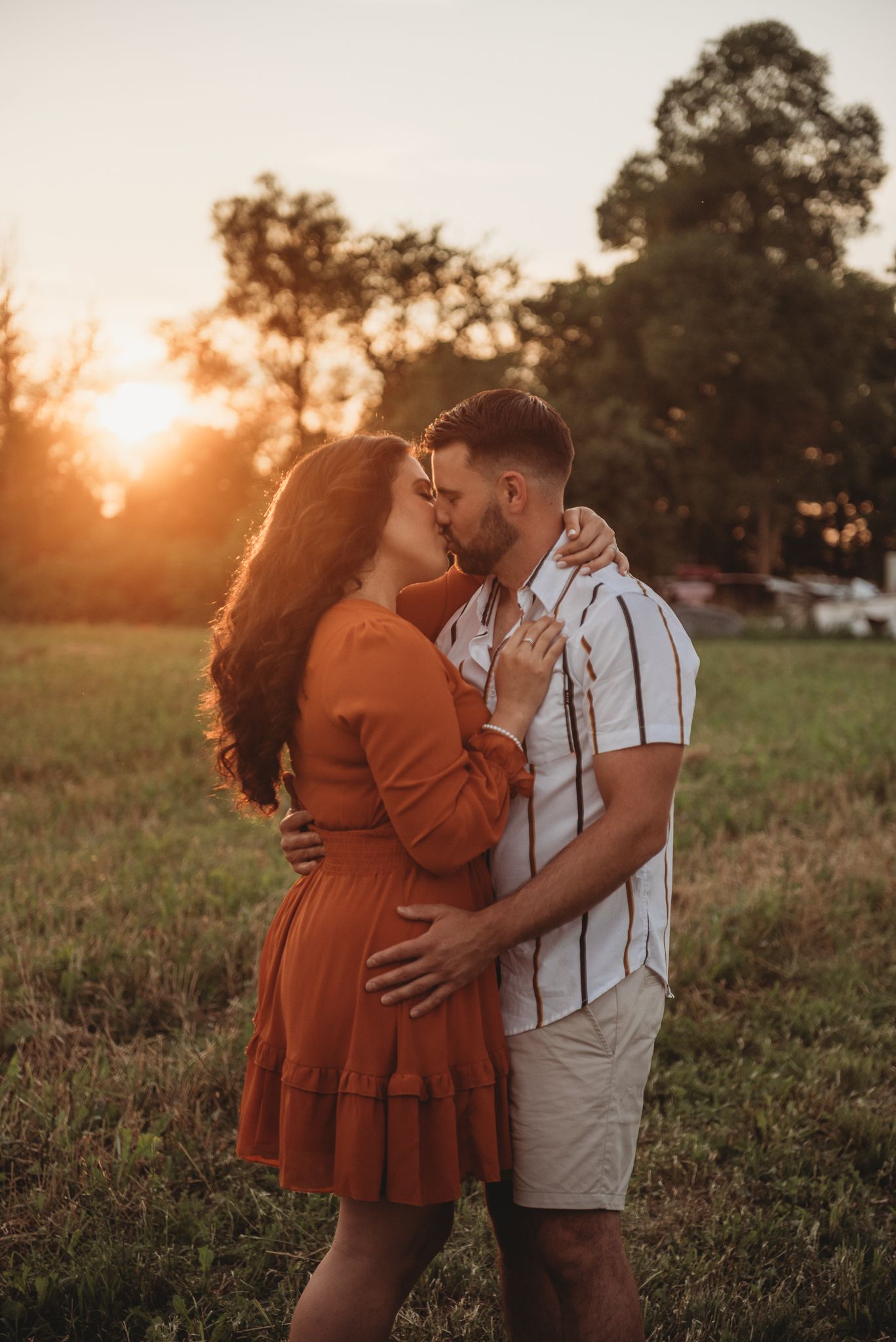 A couple shares romantic moments in a sun-drenched field at sunset, wearing rust-colored dress and white shirt.