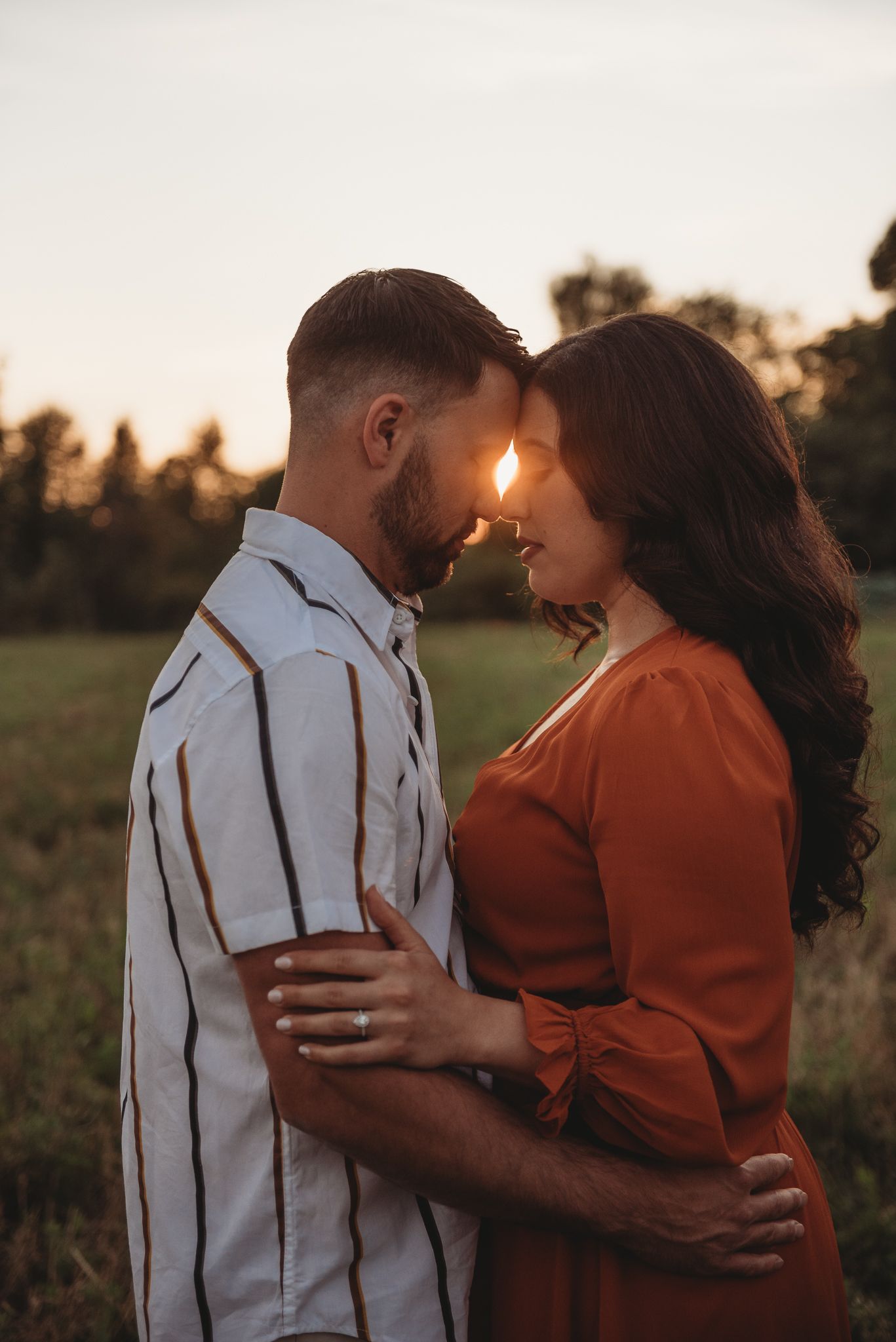 A couple embraces tenderly in a field at golden hour wearing white and rust-colored outfits.