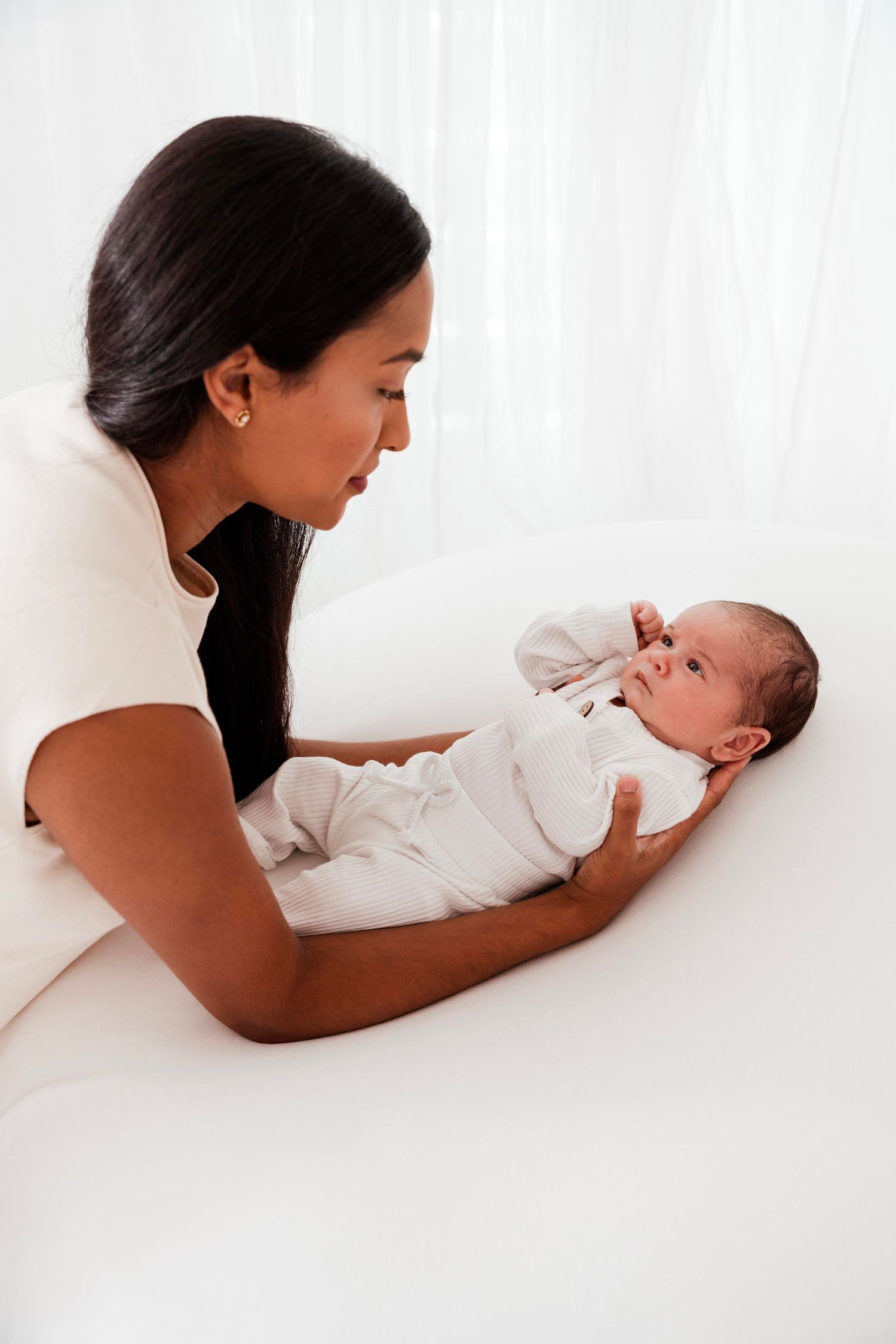 A mother lying on white bedding gazes lovingly at her newborn baby in a tender moment.