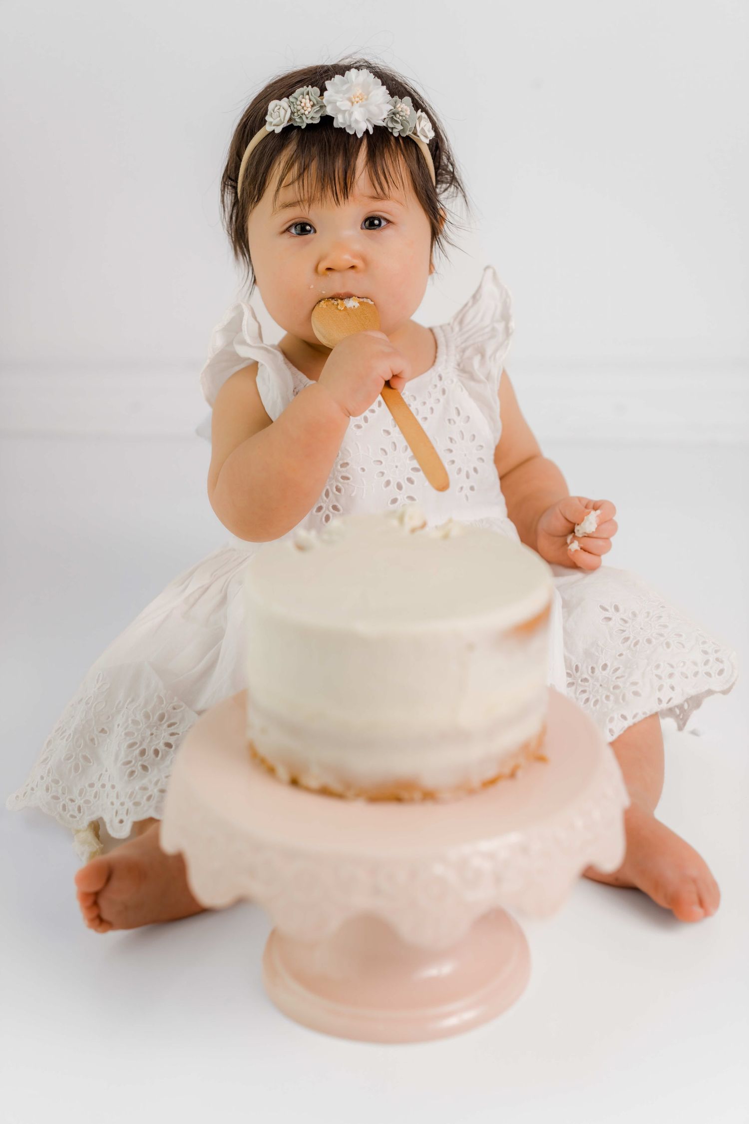 Baby in white lace dress enjoys first birthday cake smash photo session with pink and white frosted cake.