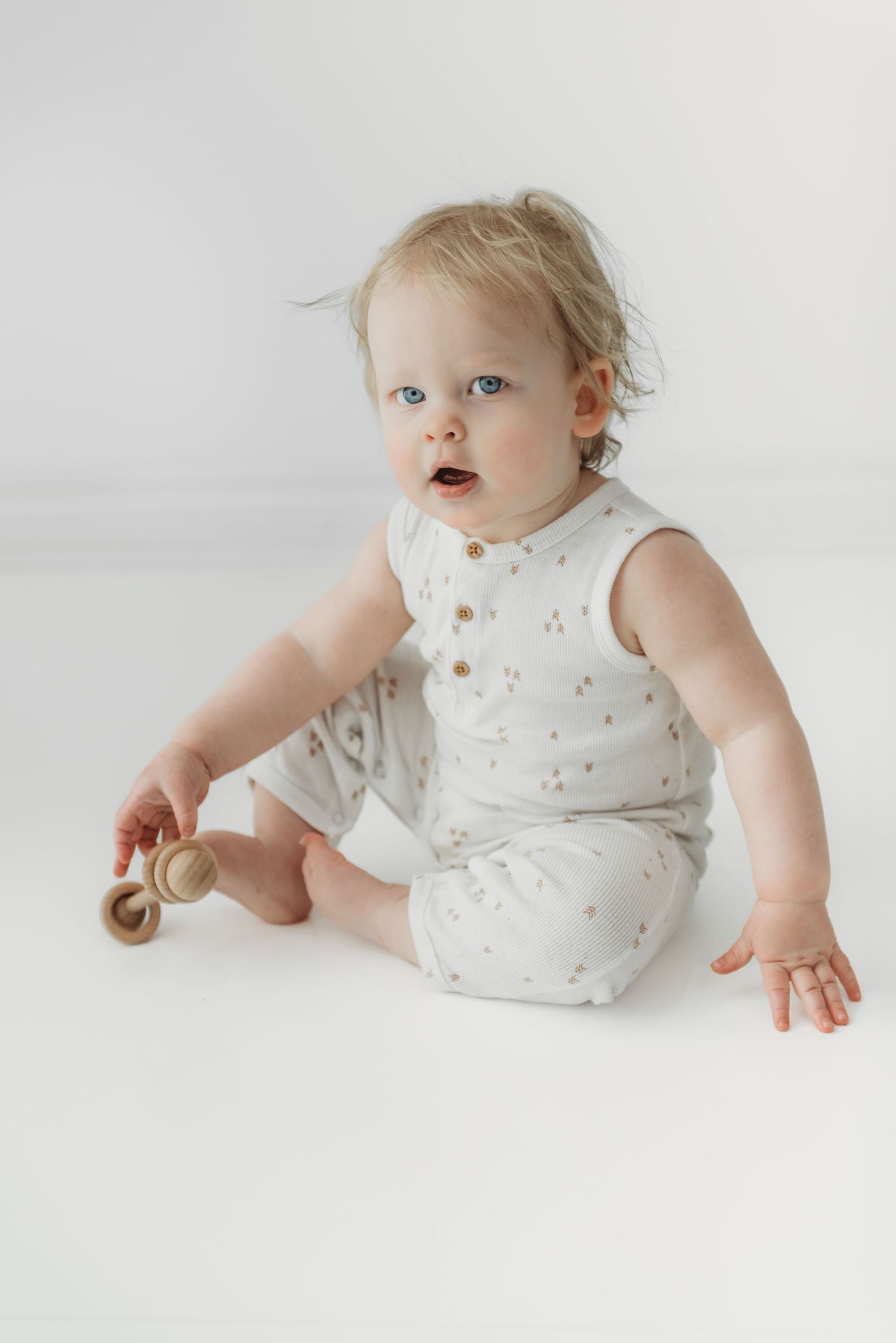 Baby in white polka dot romper sits on light background while holding wooden toy.