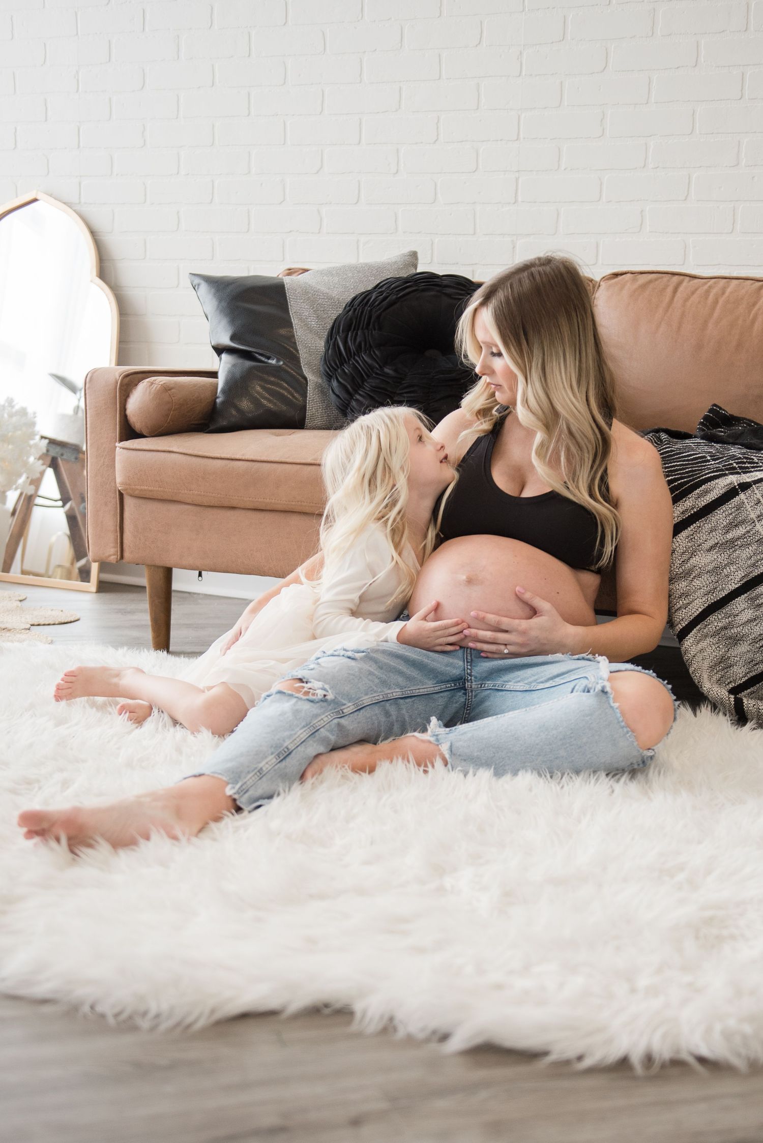 Two people resting together on a fluffy white rug beside a tan leather couch in a bright, minimalist room.
