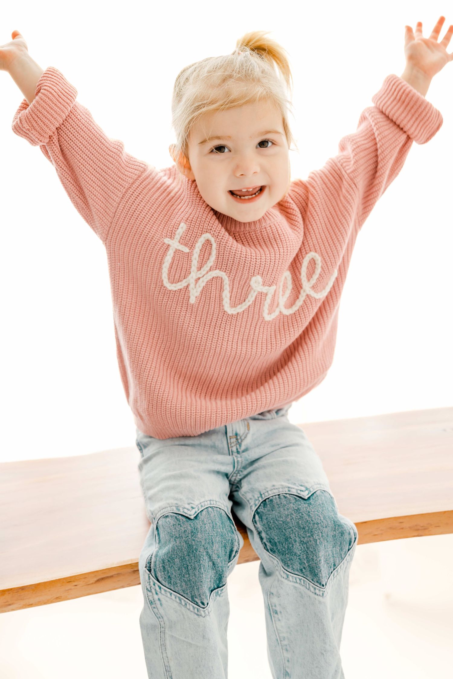 A joyful toddler wearing a pink sweater and distressed jeans jumps and plays in a bright indoor setting.