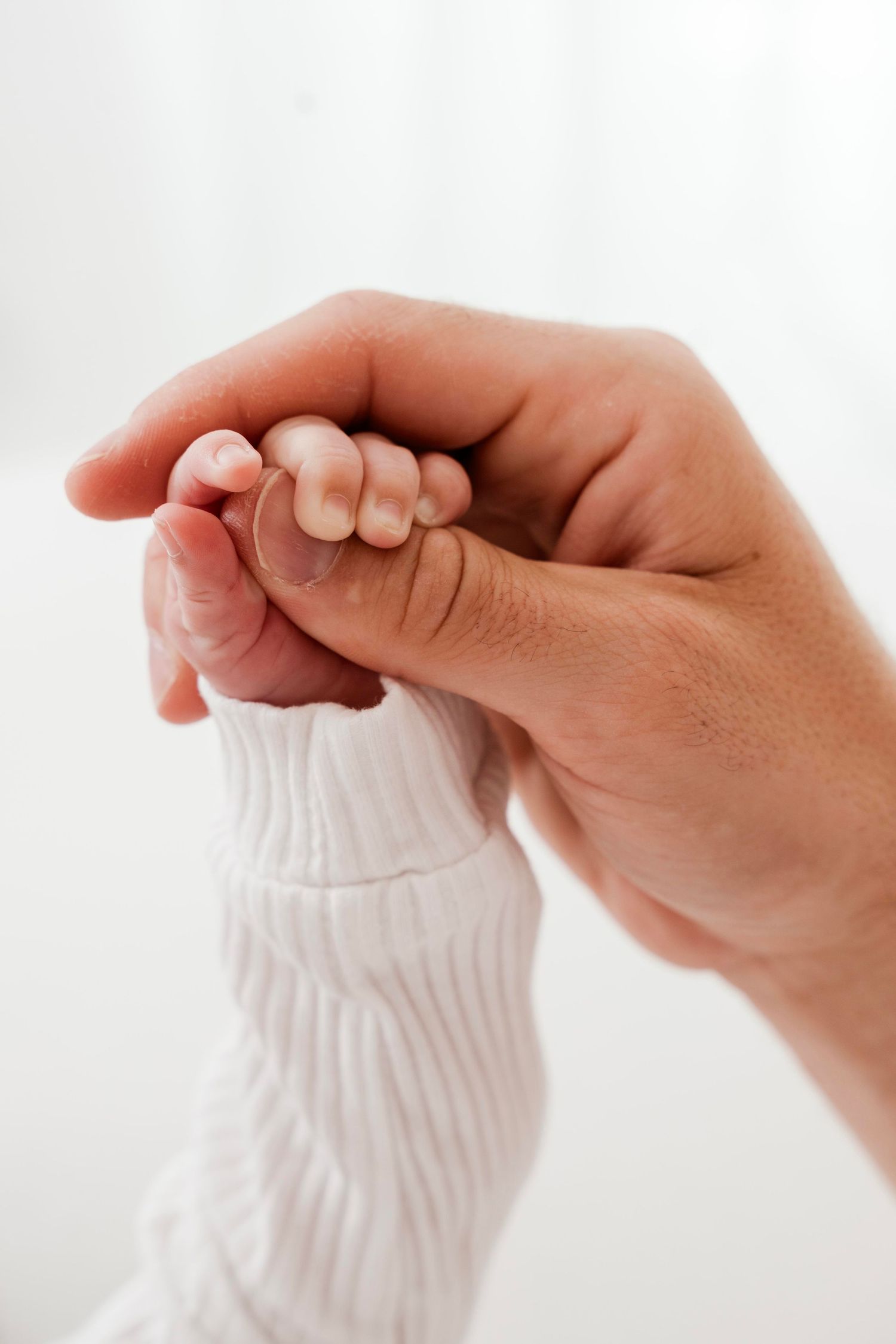 A tender moment captured as an adult hand gently holds a tiny baby's hand against a white background.