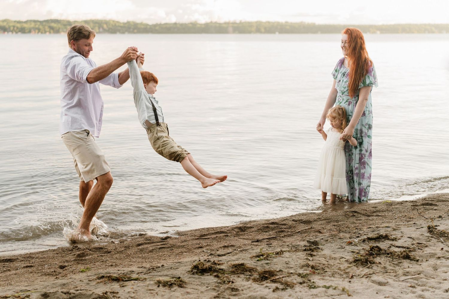 Family enjoying a sunny beach day, father swinging young boy over water while mother stands with daughter by the shore.