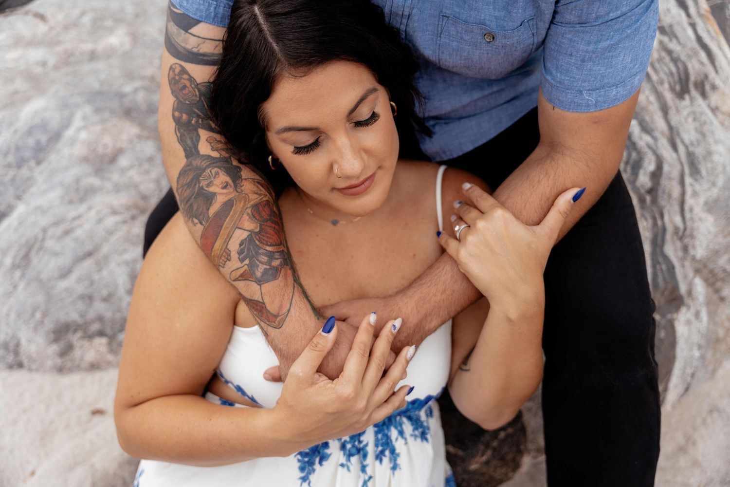 A couple embraces on a rocky surface, with one person wearing a white and blue dress.