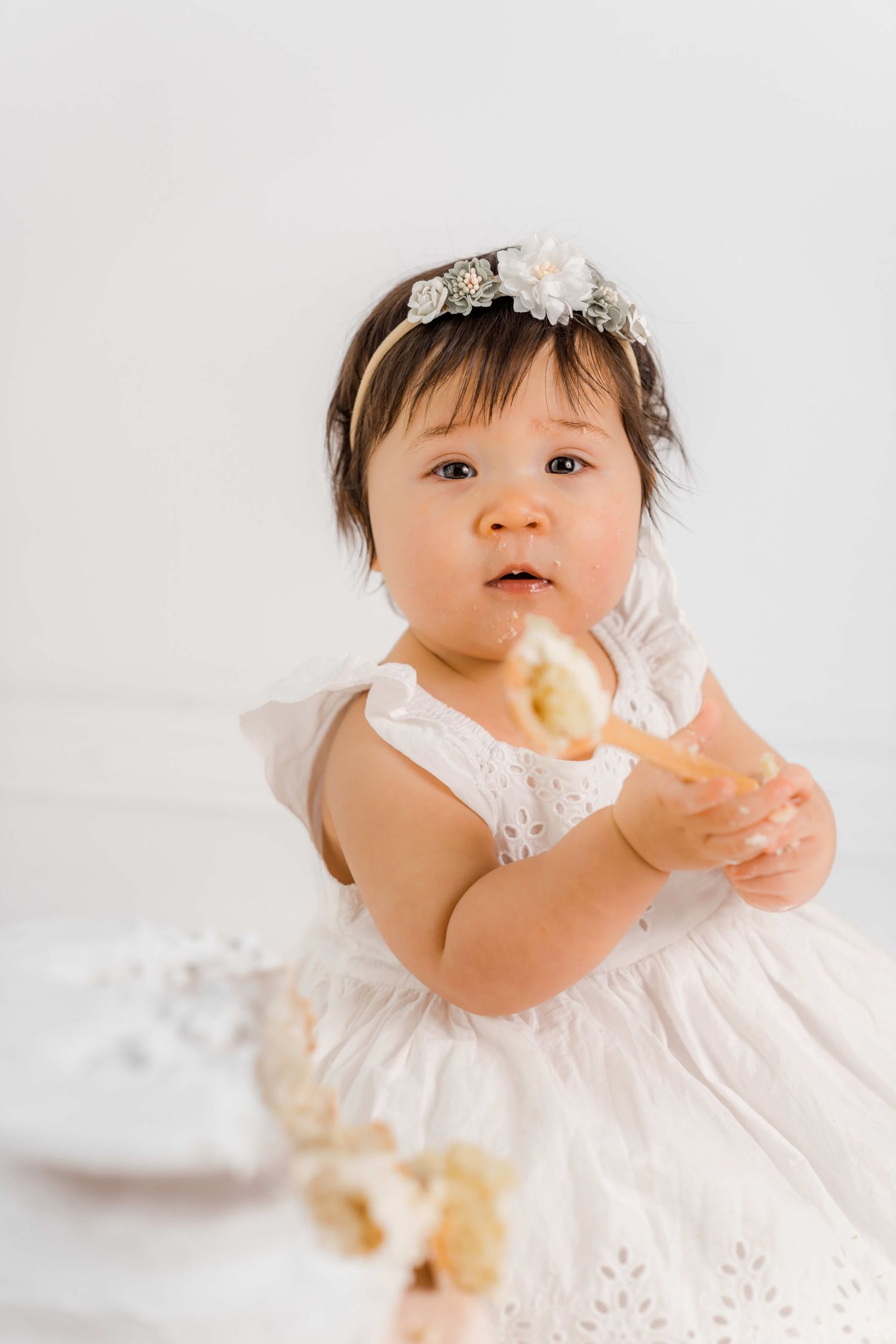 Baby in white lace dress crawls and plays with a toy while wearing a delicate floral headband.