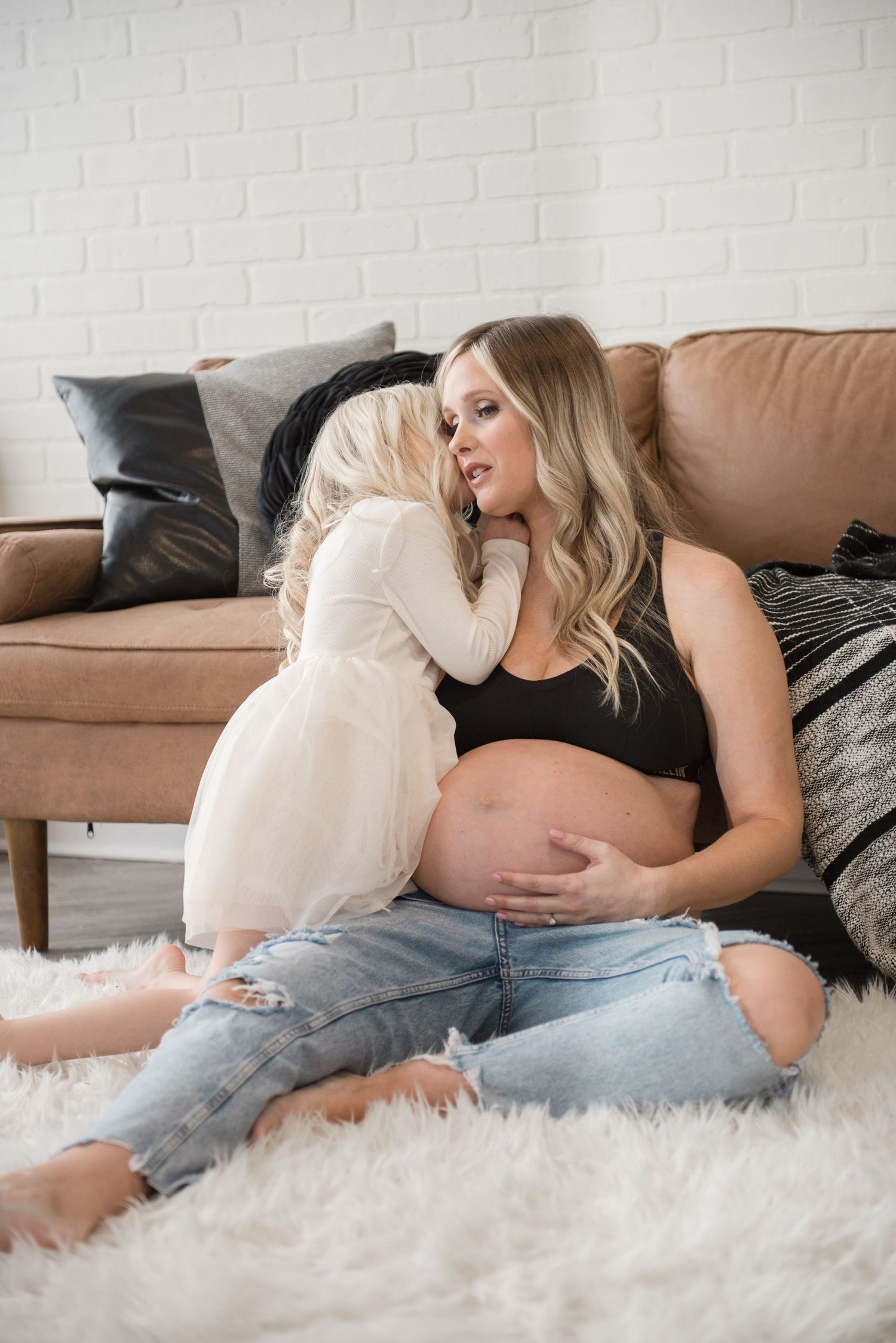 A pregnant woman in ripped jeans and black crop top sits on a plush white rug with a small dog beside her.