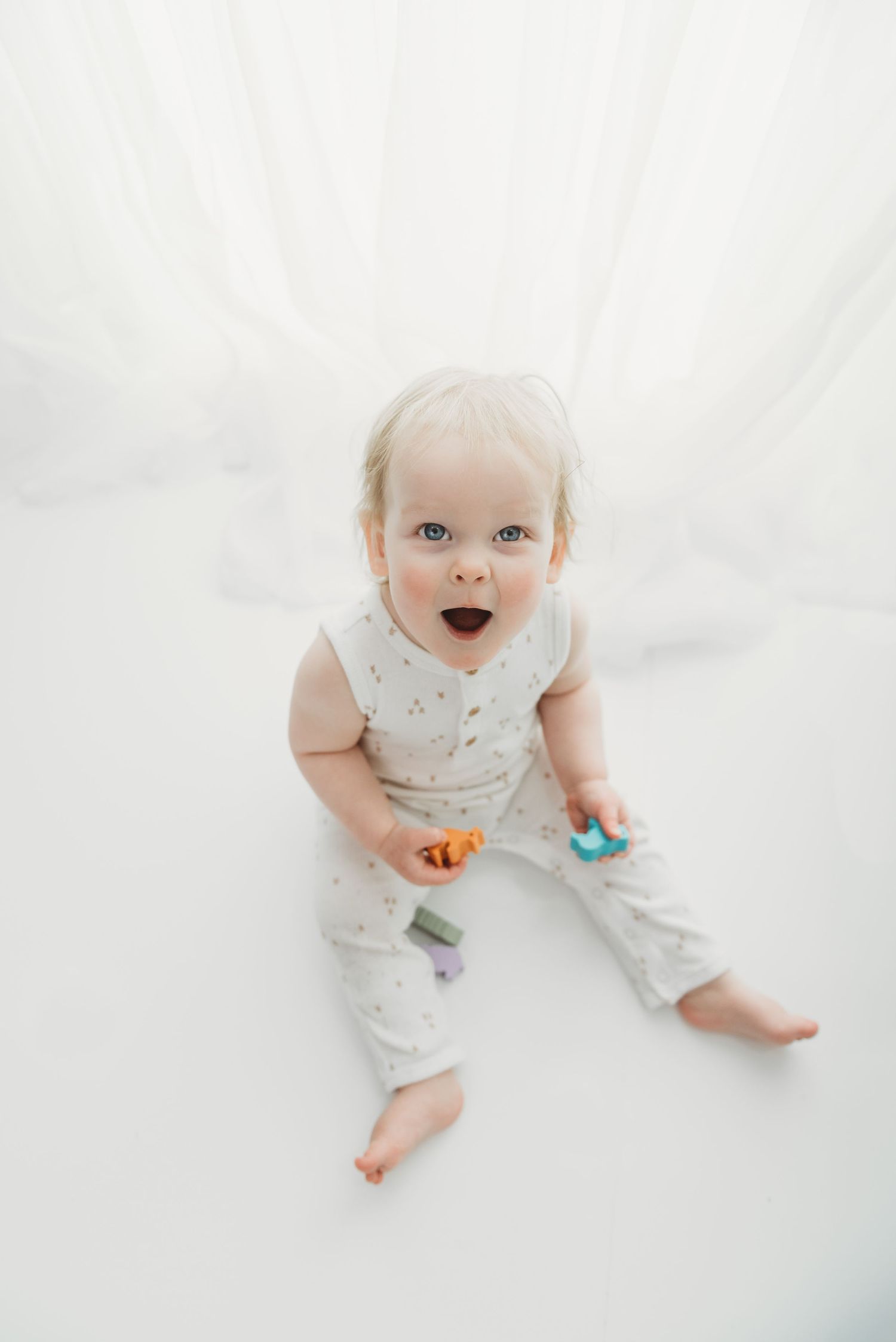 Baby in white polka dot outfit lying on white blanket holding colorful toys and smiling with joy.