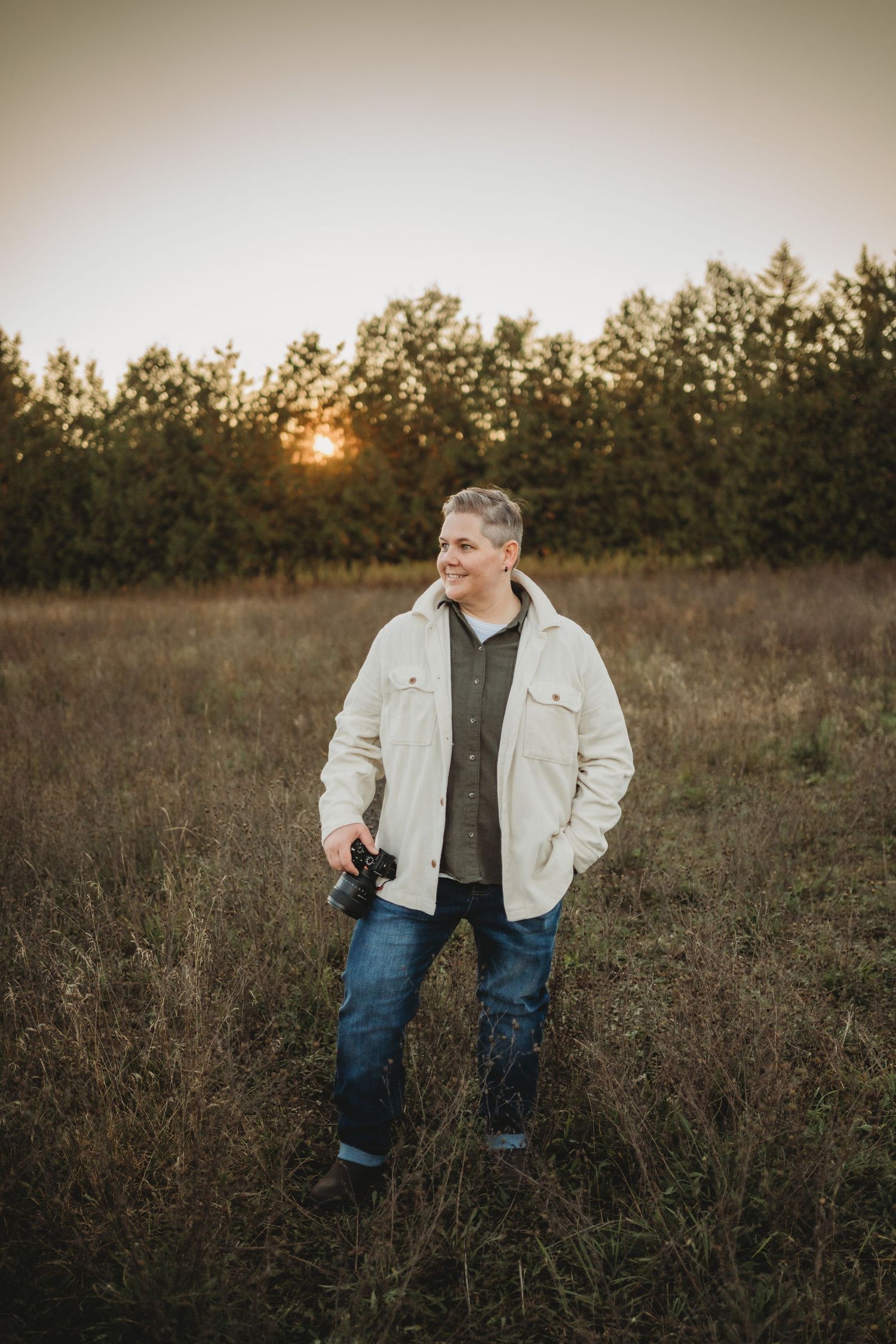 A person in a light denim jacket and jeans stands in a golden field at sunset while holding a camera.