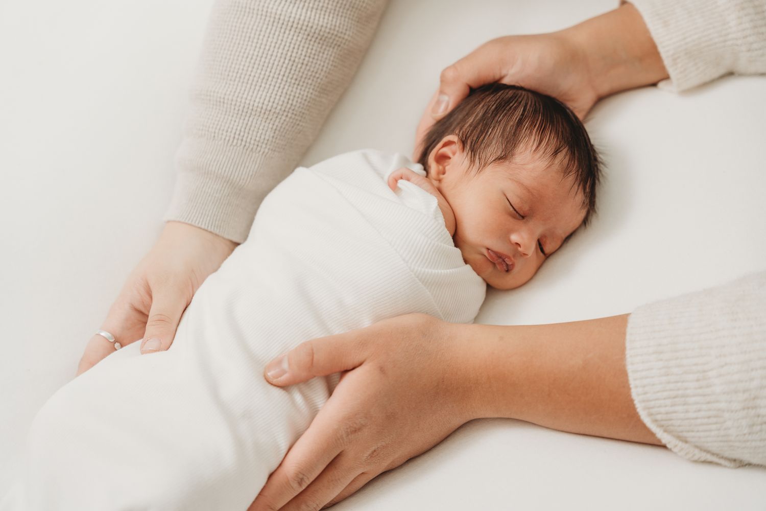 A sleeping baby in white clothing lies peacefully on white bedding while a gentle hand provides comfort.