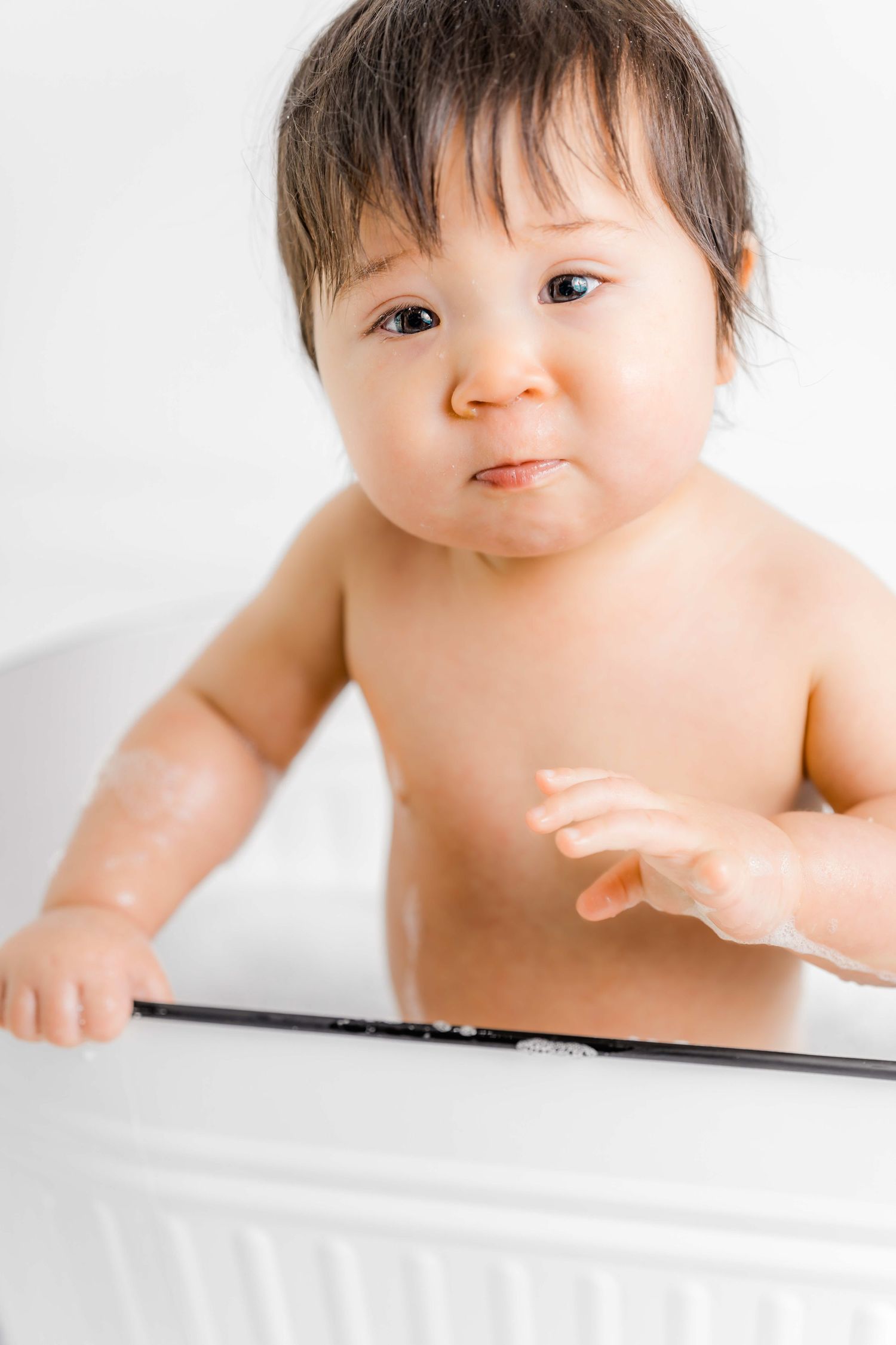 Baby with dark hair leans over white surface while reaching forward with bare shoulders against light background.