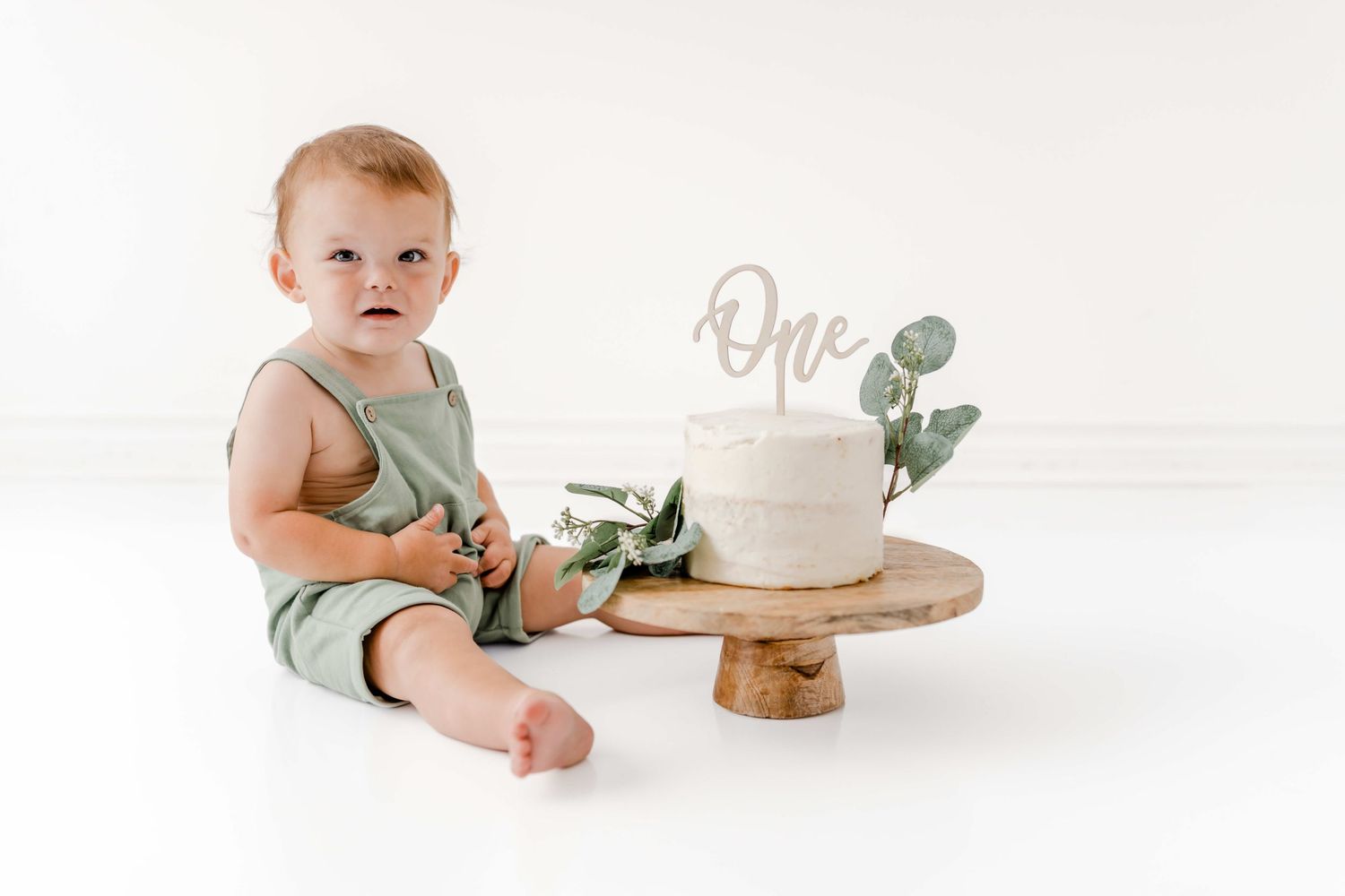 Baby in green outfit sits near wooden cake stand with white cake and eucalyptus leaves in bright white setting.
