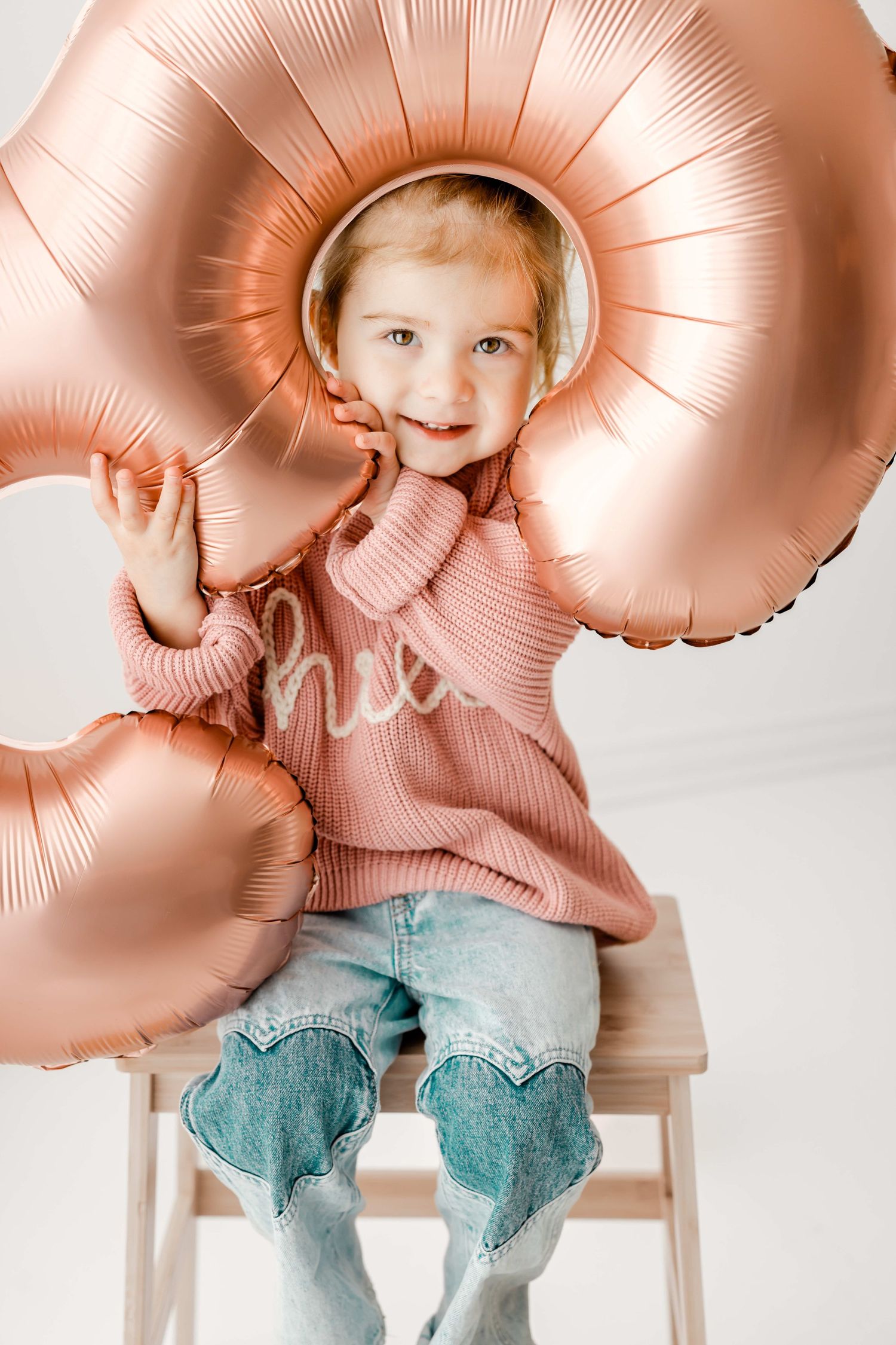Child in pink sweater sitting with rose gold heart-shaped balloons against white backdrop.