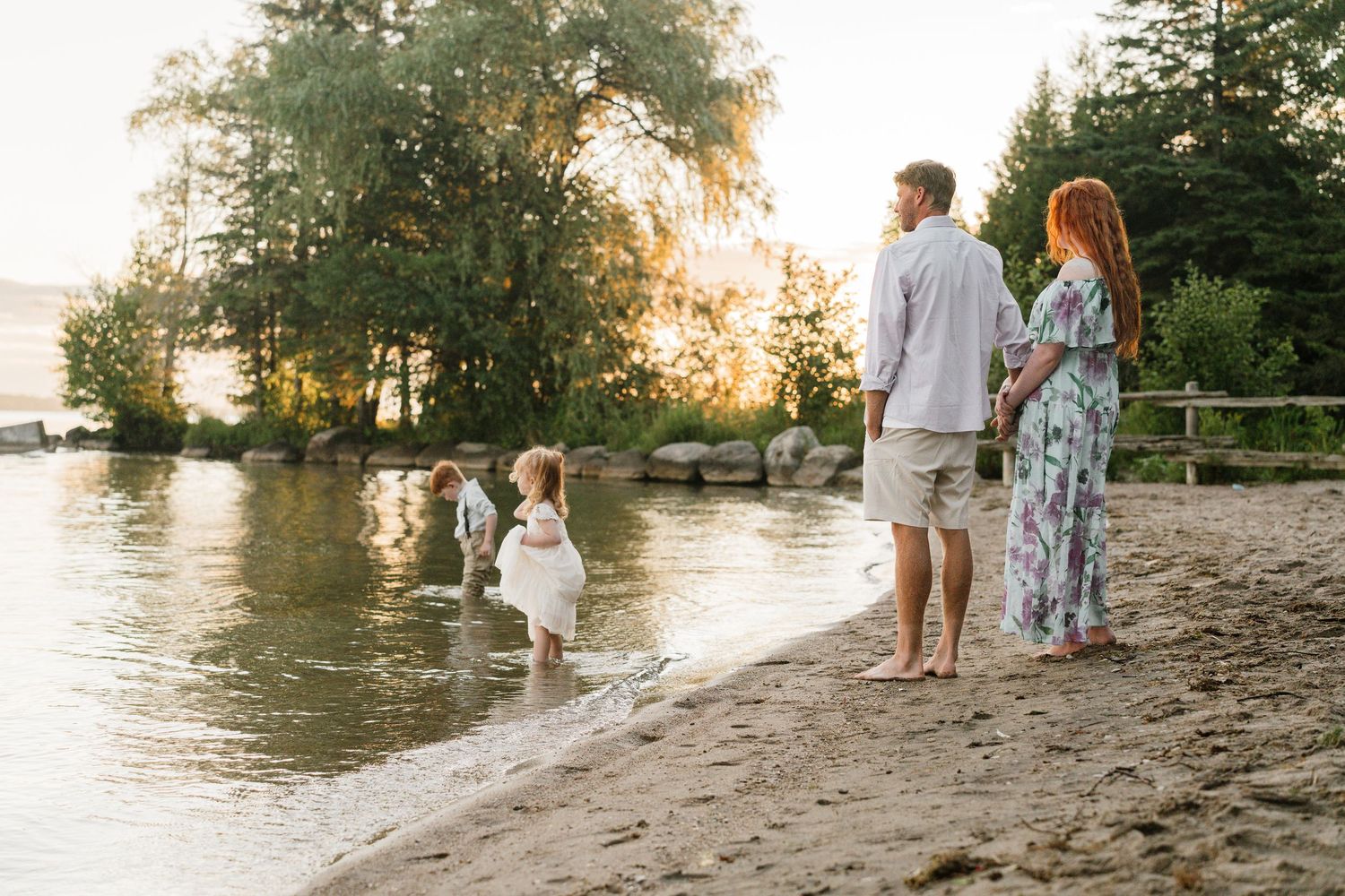 Family enjoying a sunset beach walk, with two young children playing at the water's edge near lush green trees.
