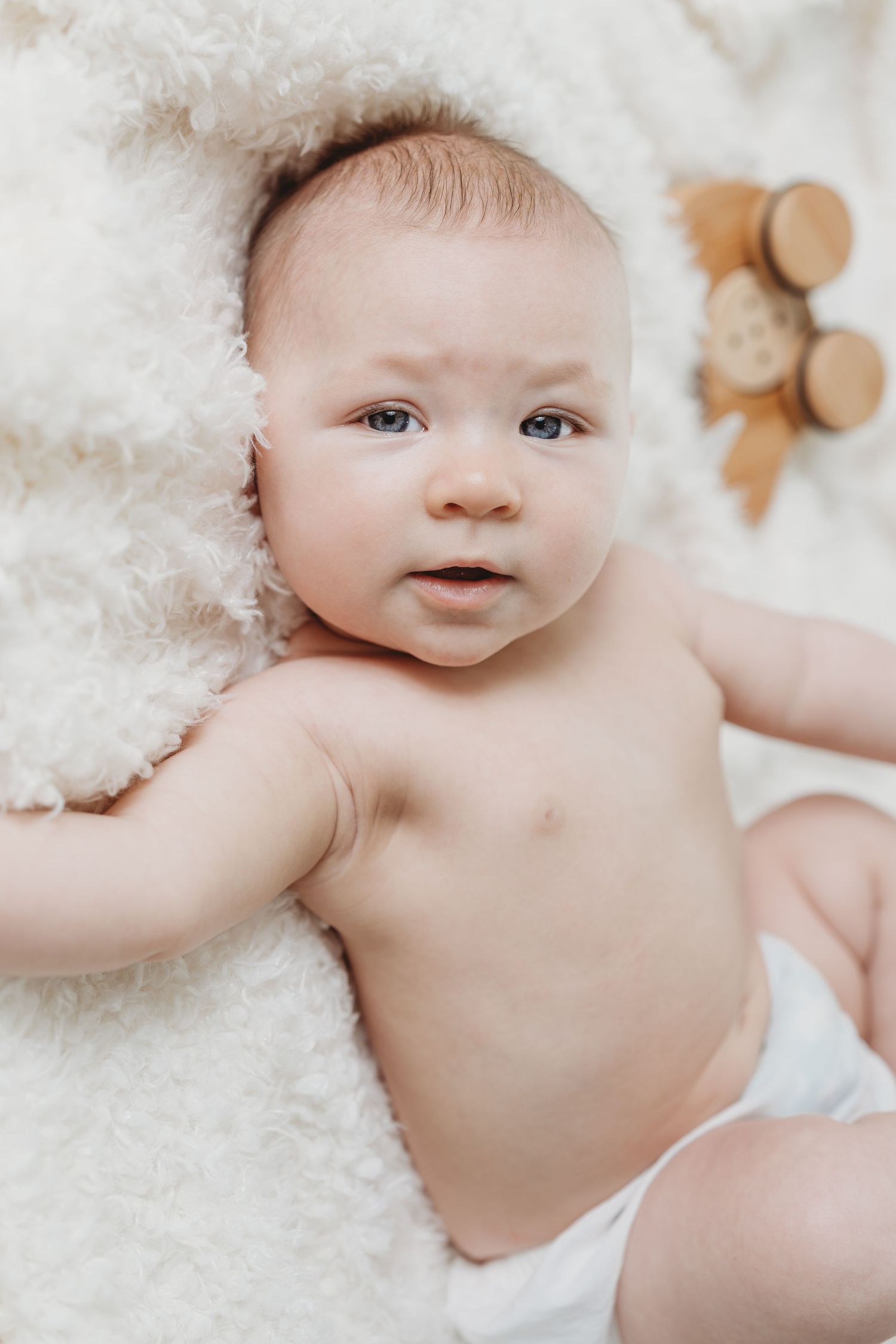 A sweet baby lies on white bedding with a plush teddy bear in the background.