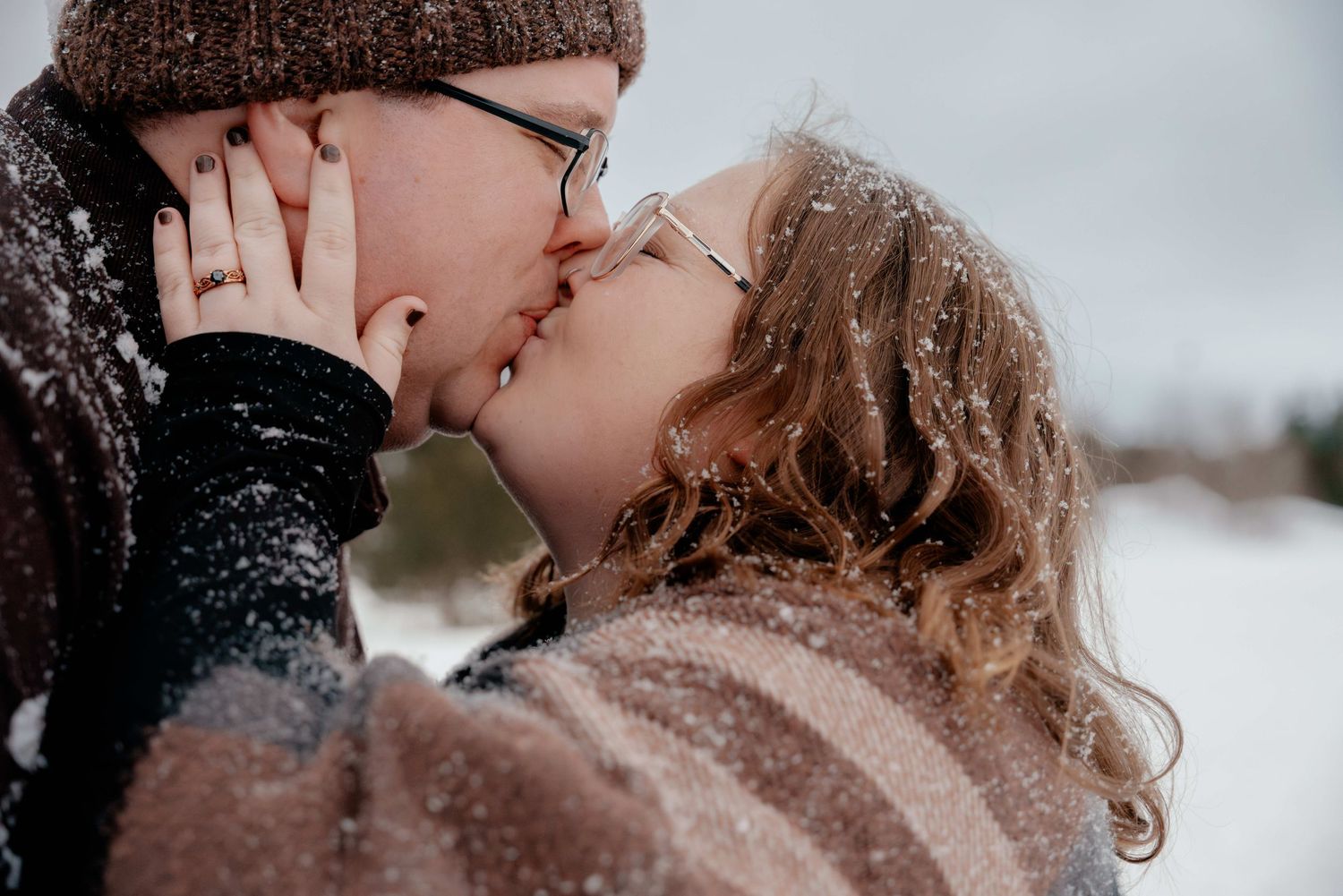 A romantic couple shares a tender kiss in a snowy winter setting with snowflakes falling around them.