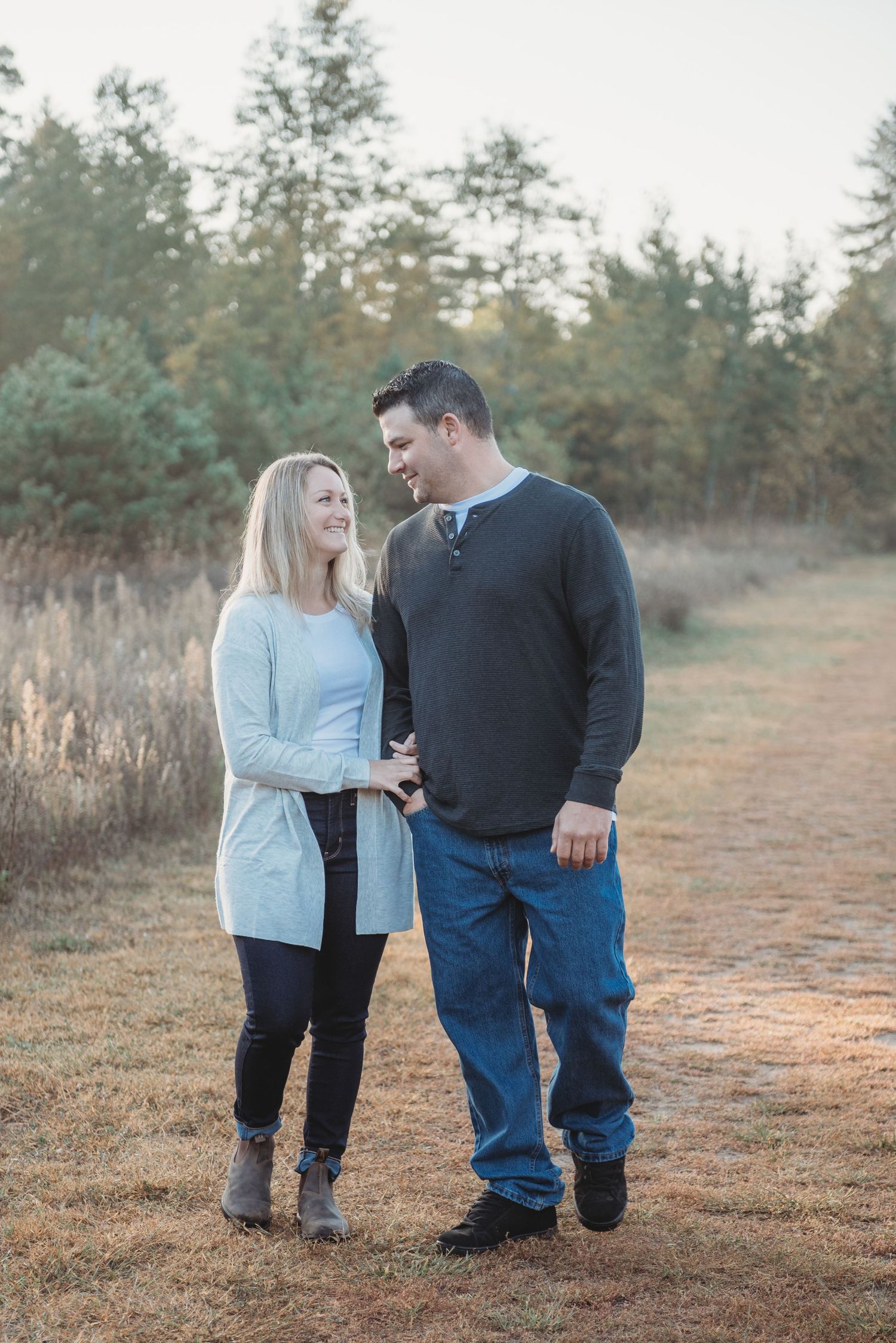 A couple stands together on a dirt path surrounded by evergreen trees during a misty autumn afternoon.