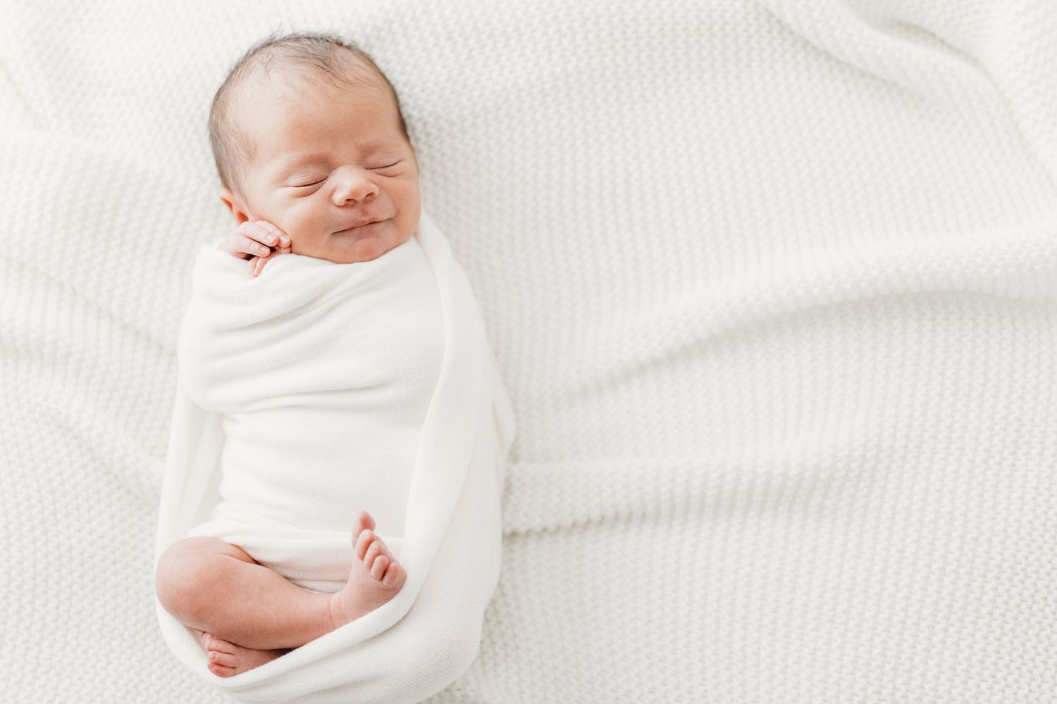 Newborn baby sleeps peacefully wrapped in white blanket on textured white background.