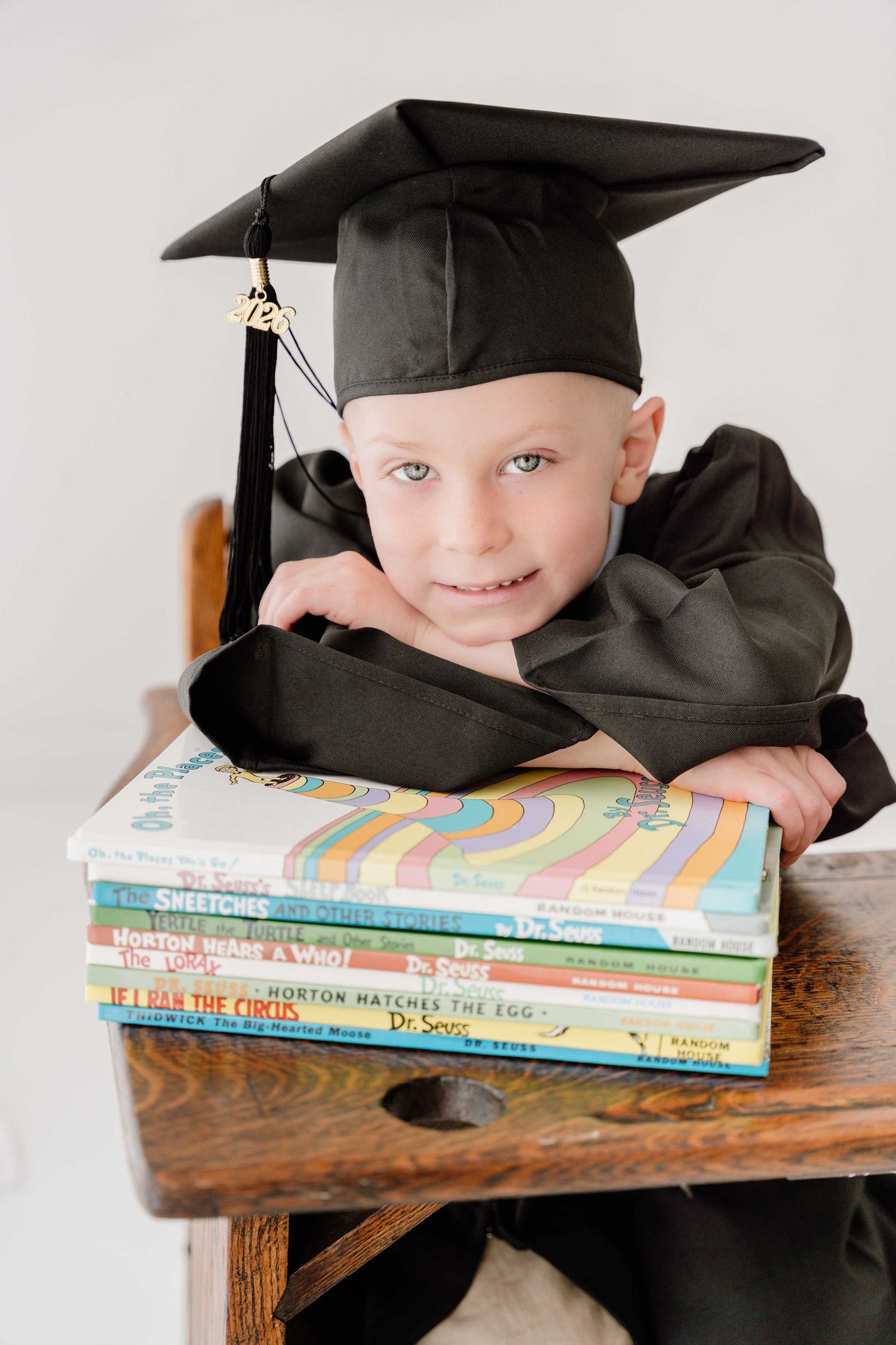 Young child in black graduation cap and gown smiling while leaning on stack of Dr. Seuss books at wooden desk.
