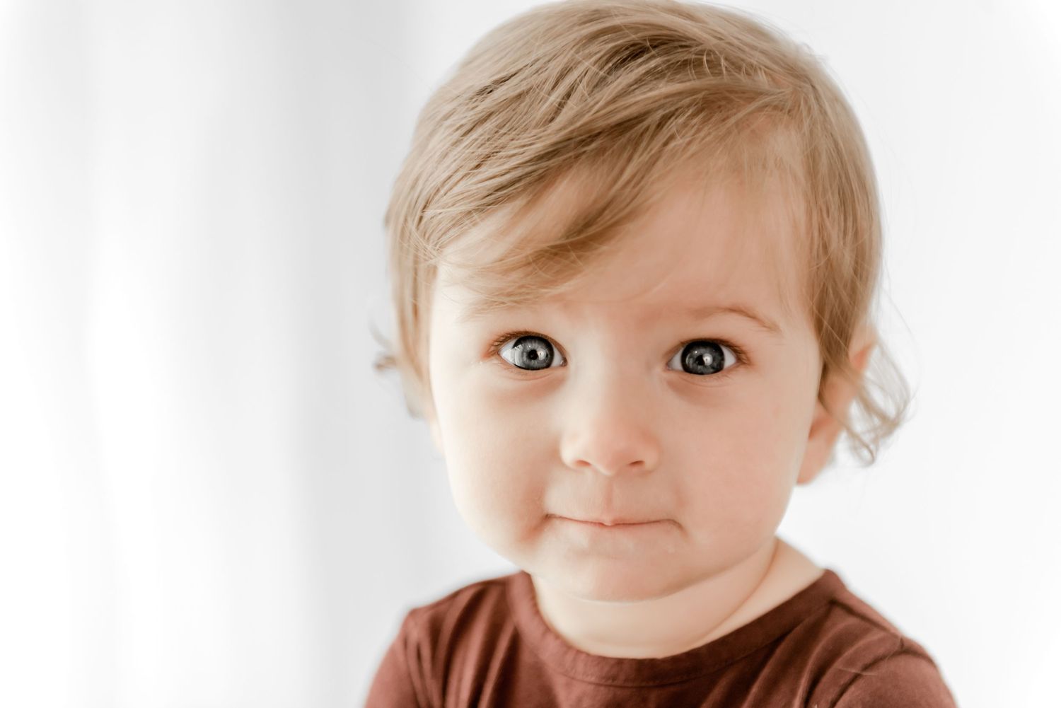 A young child with light brown hair wearing a brown shirt looks directly at the camera against a white background.