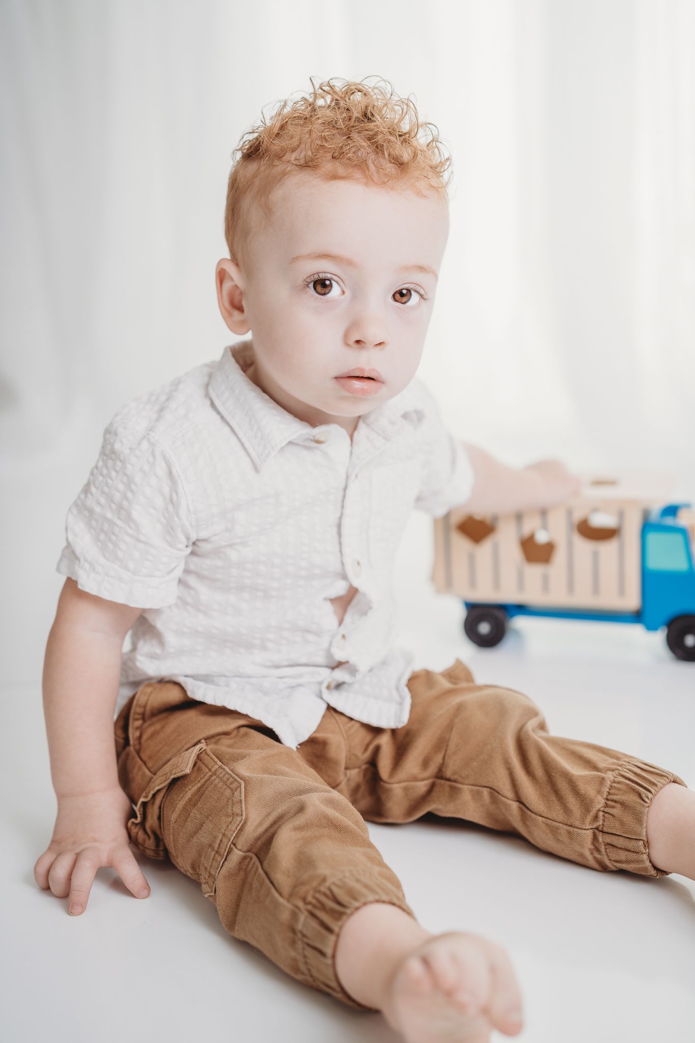 A small child in a white shirt and brown pants lies on a white background with a blue toy nearby.