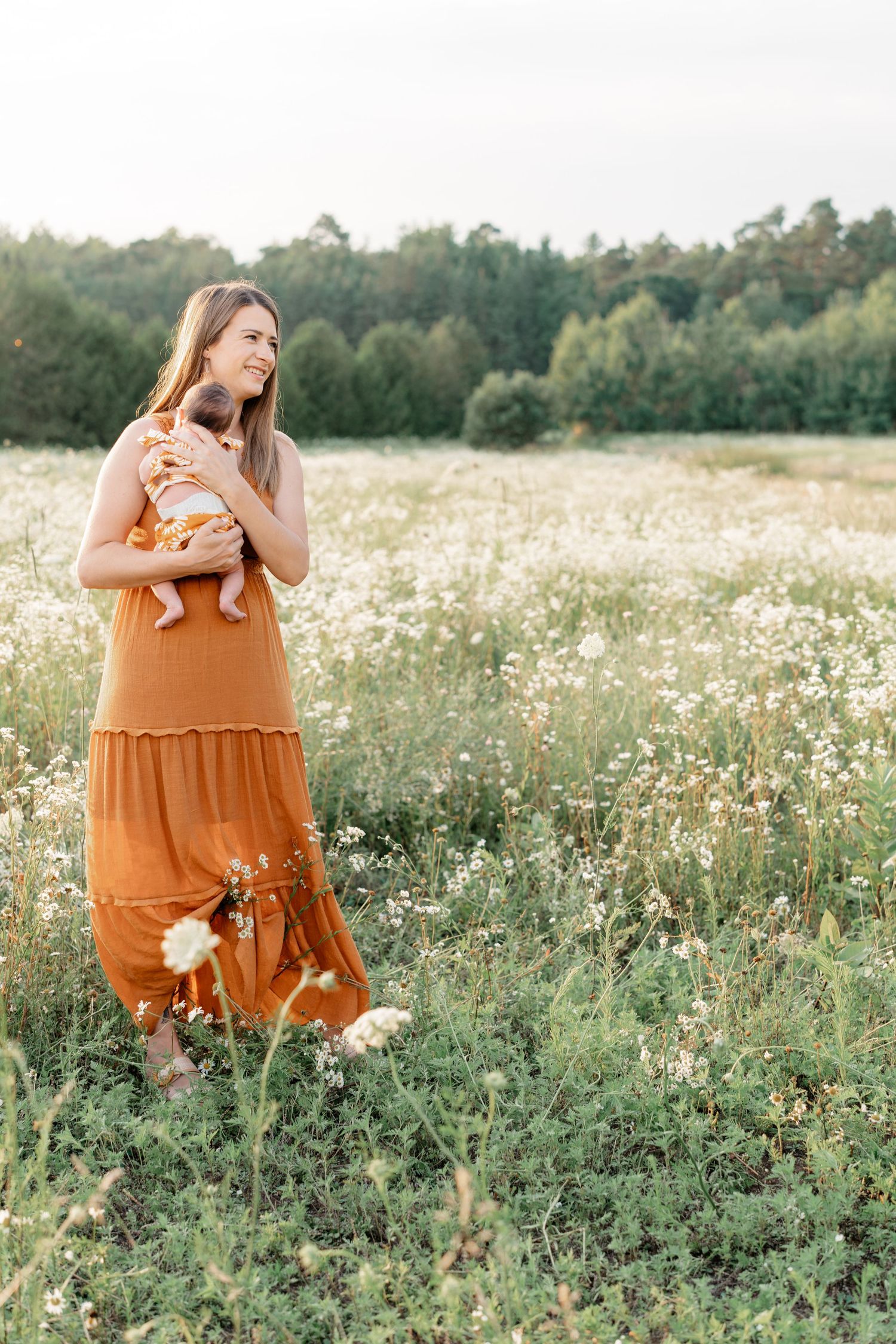 Woman in flowing orange dress stands in a wildflower meadow at golden hour with trees in the background.