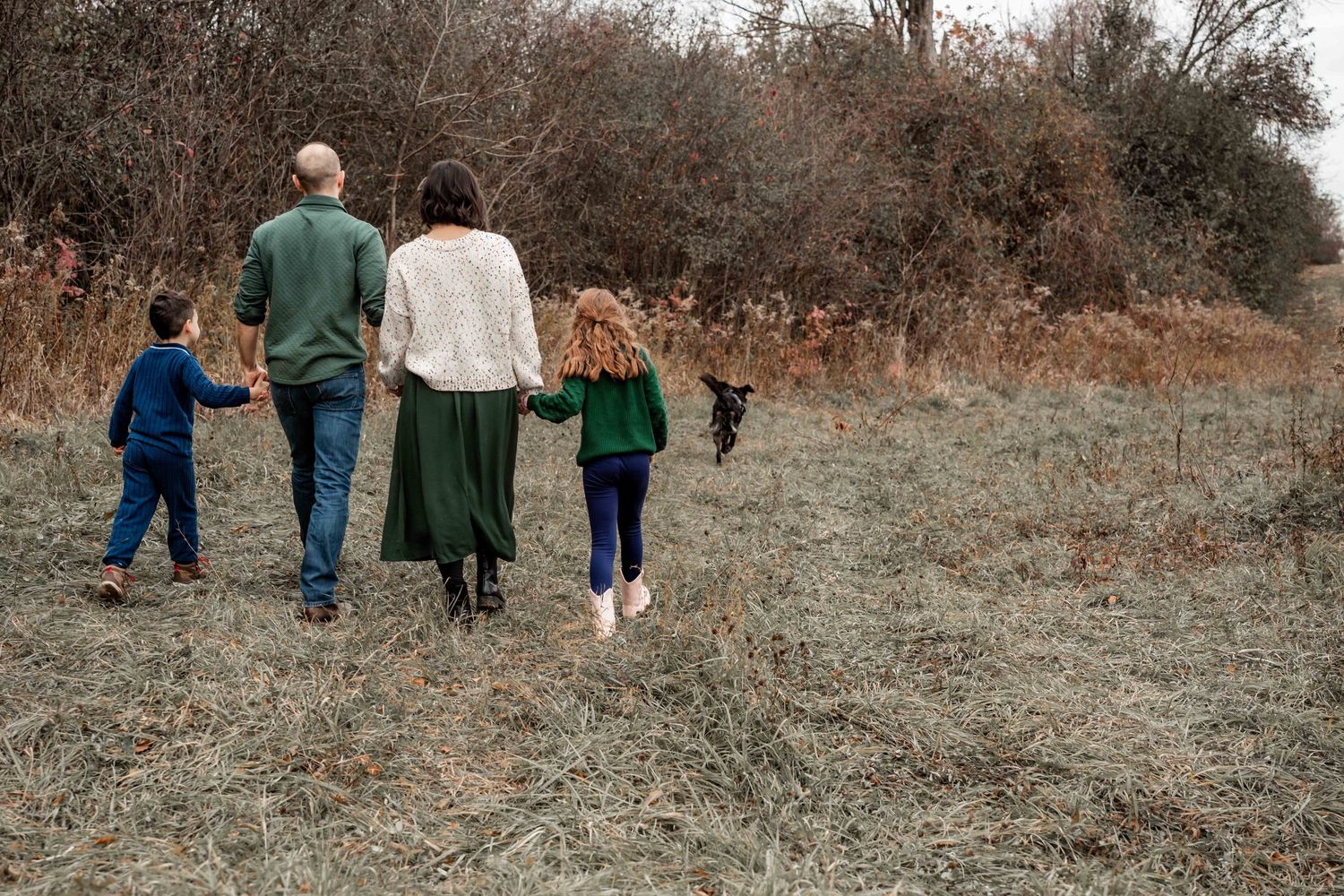Family walking together with their dog on a rustic path through autumn brush and dried grass.
