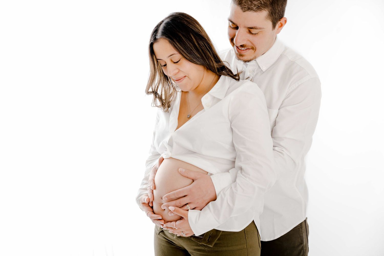 A couple in white shirts embraces during a maternity photo session against a bright white background.