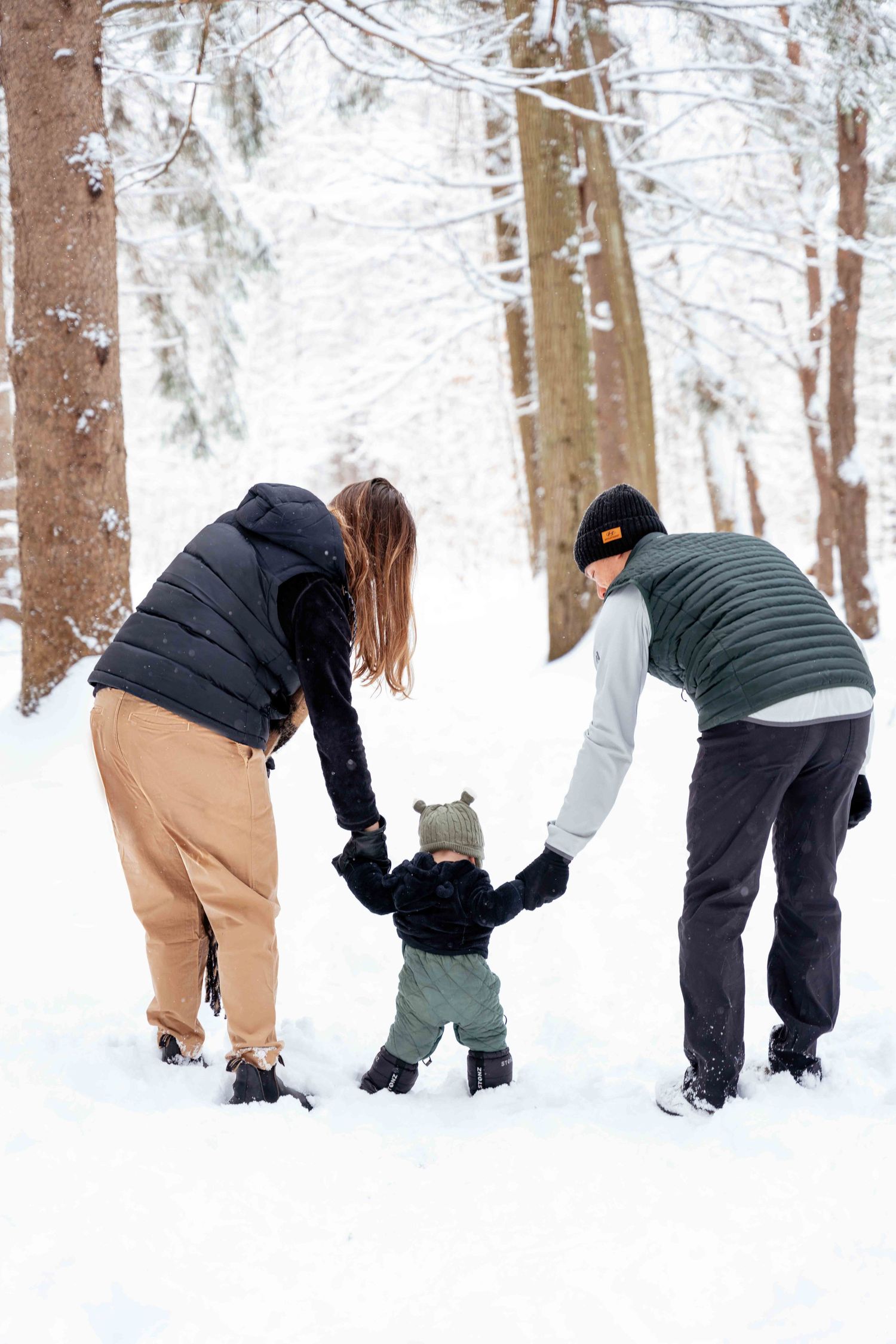 Two adults help a small child learn to walk in a snowy winter forest setting.