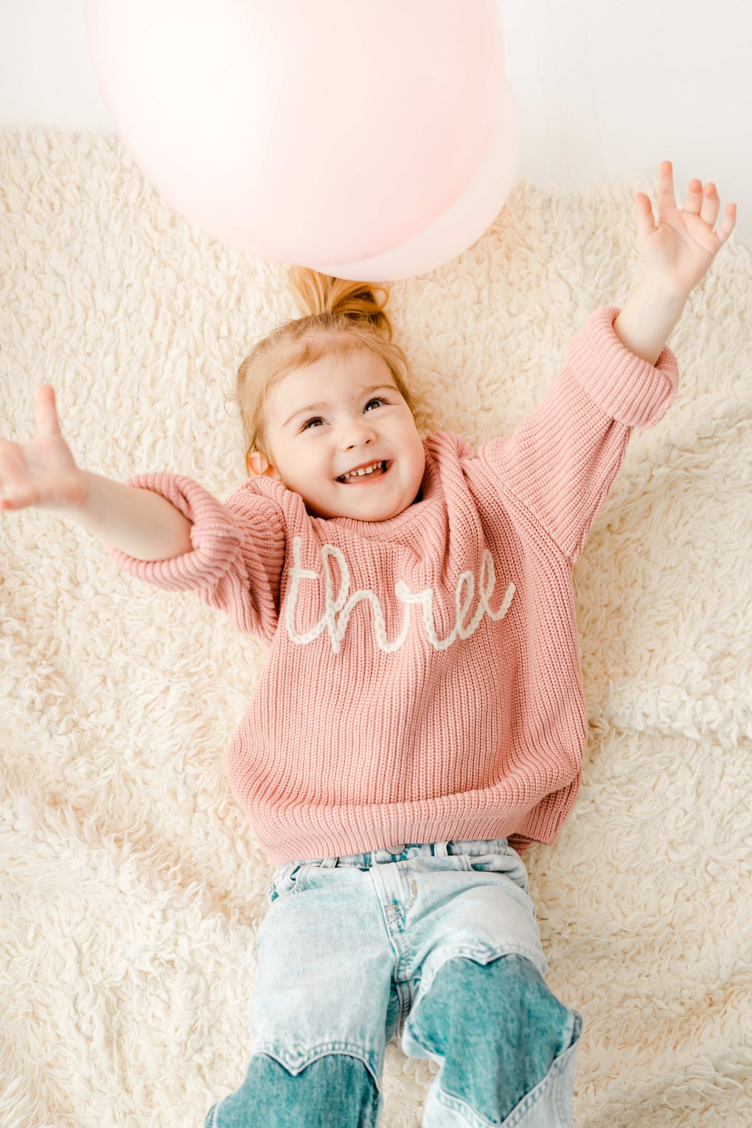 A joyful toddler in a pink sweater and jeans lies on a cream carpet with pink balloons in the background.