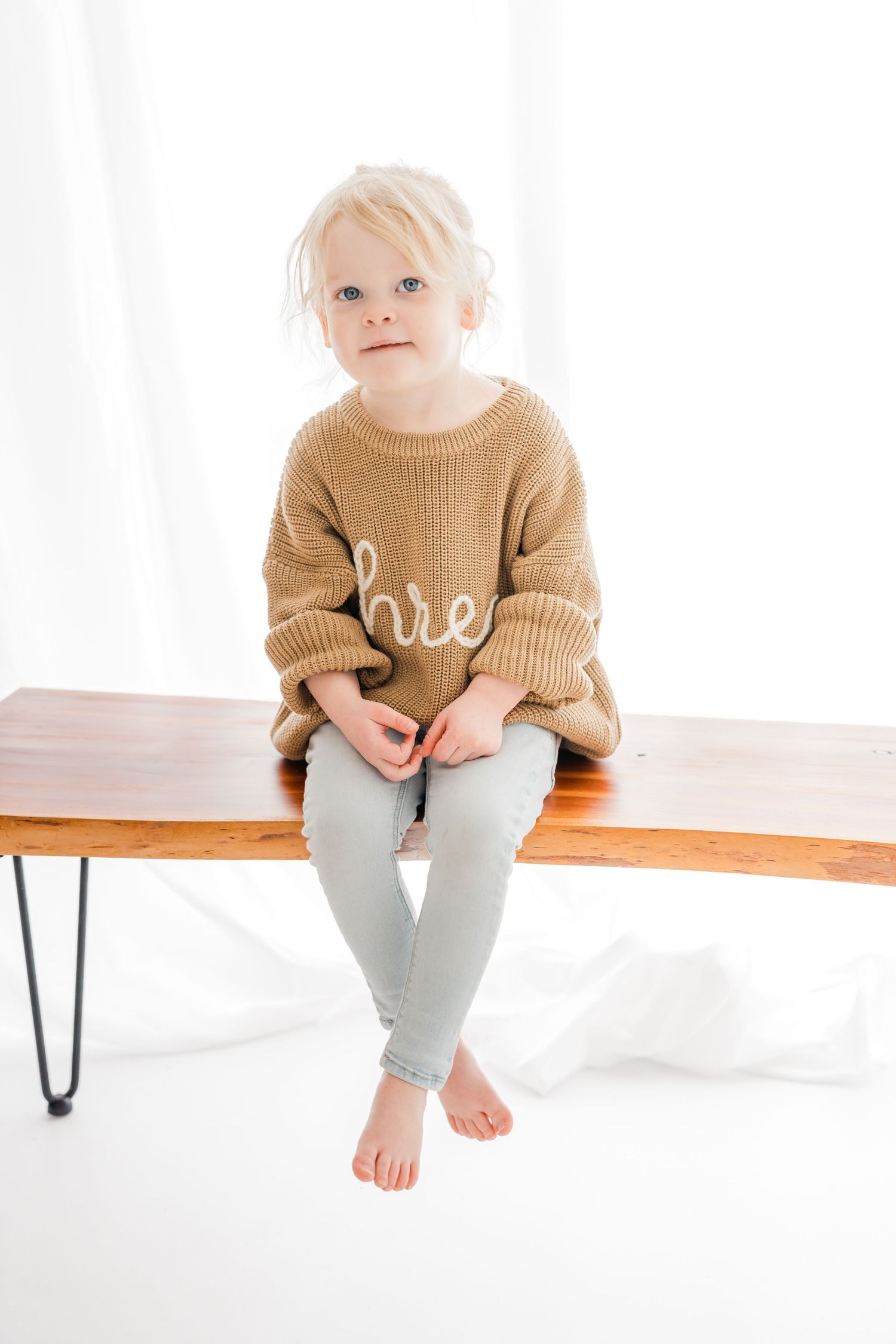 Small child wearing a tan sweater and gray pants sits on a wooden bench against white background.