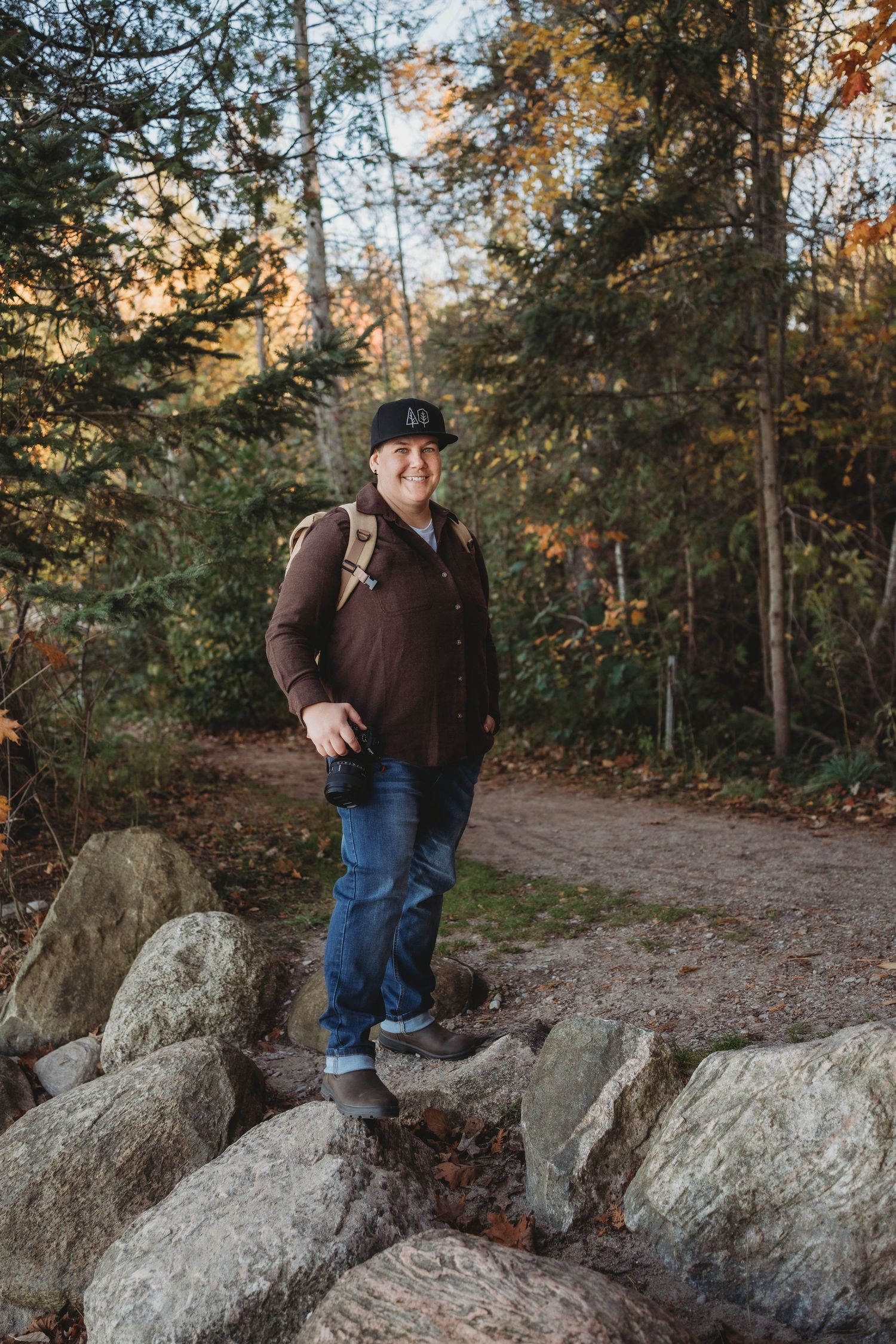 Person in brown jacket and jeans stands on large granite boulders along a wooded autumn trail at sunset.