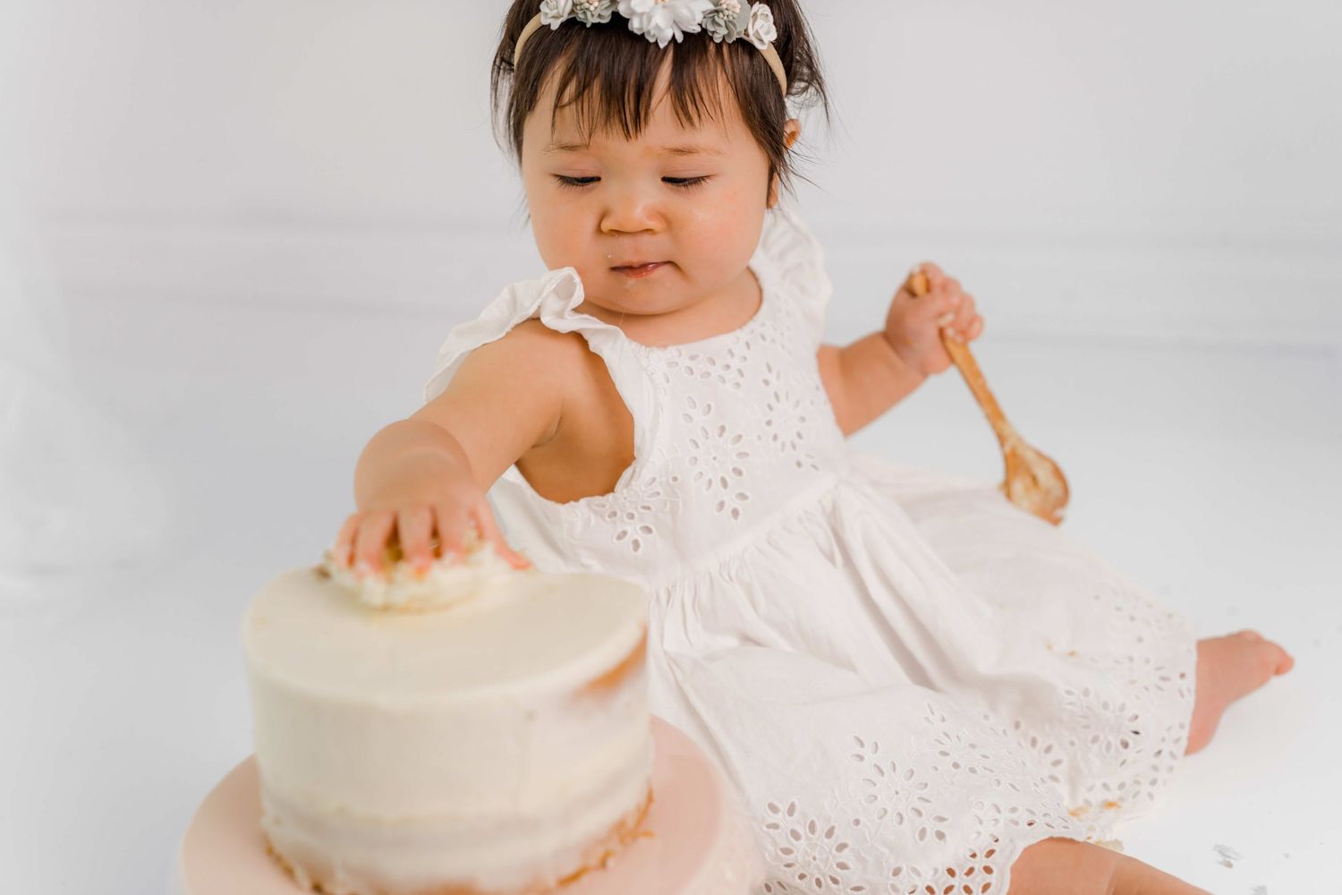 Baby in white eyelet dress reaches for white frosted birthday cake during first birthday photoshoot.