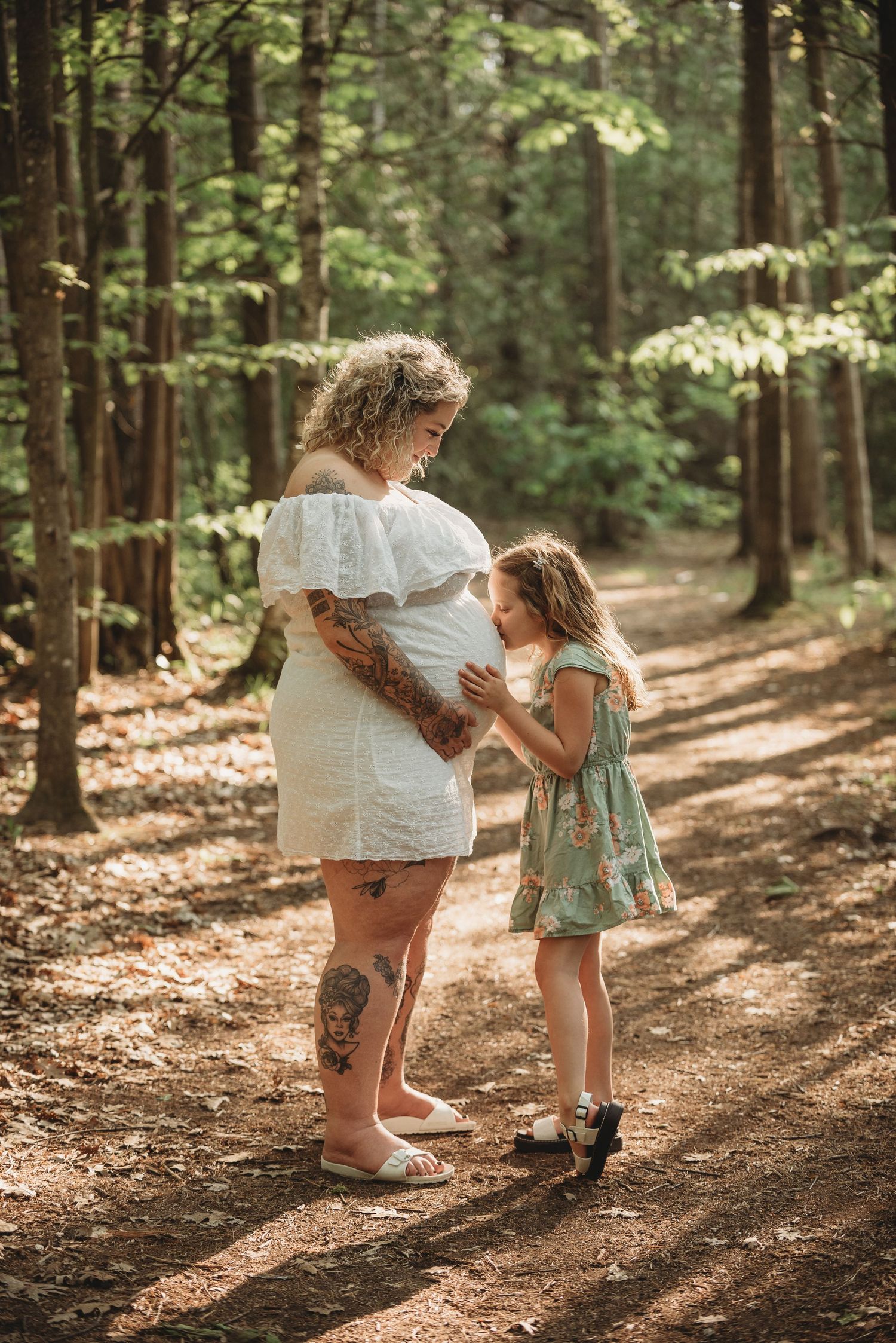 A tender moment between parent and child on a sunlit forest path surrounded by tall trees and fallen leaves.