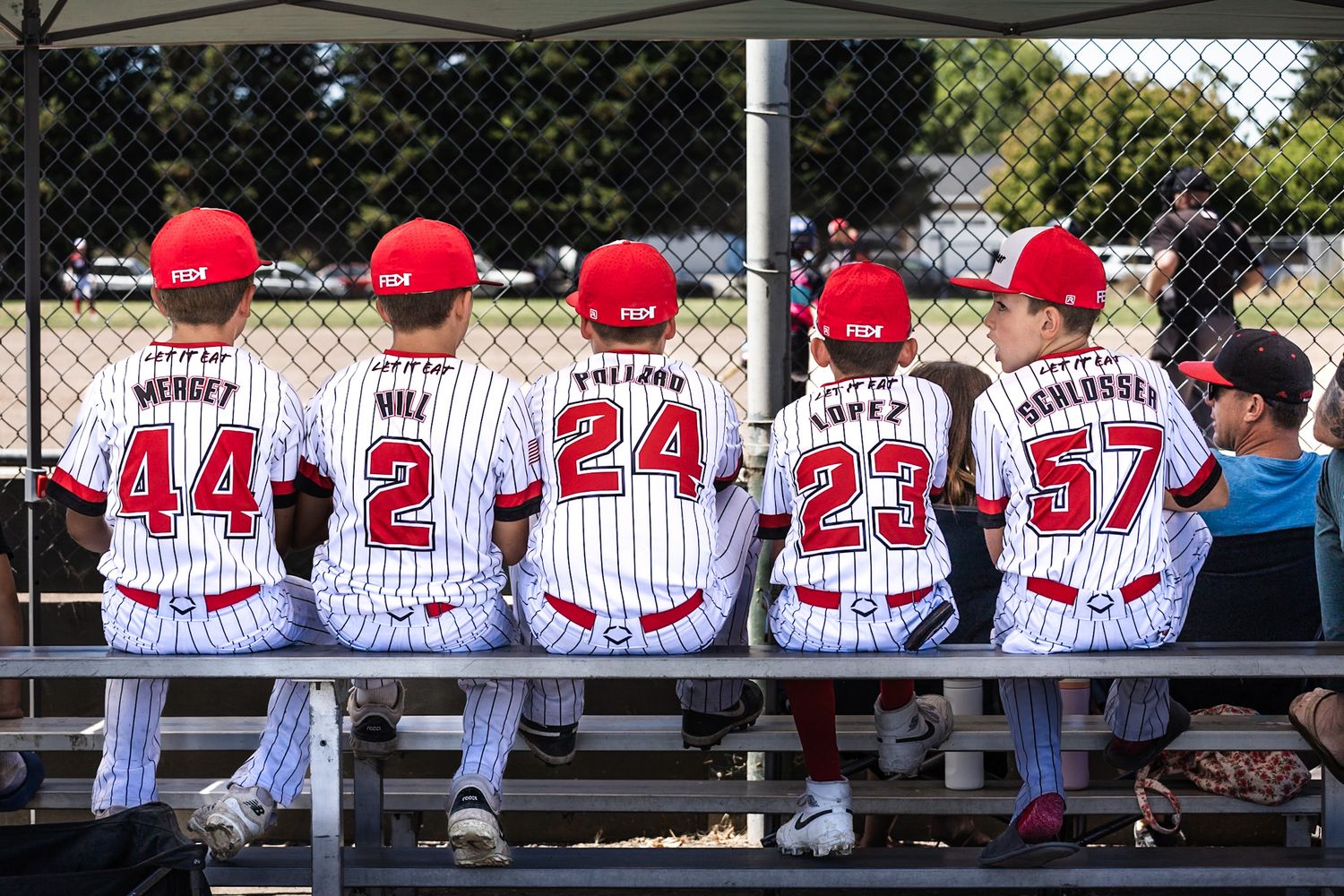 Five young baseball players sit on a dugout bench wearing red helmets and numbered white pinstripe jerseys.