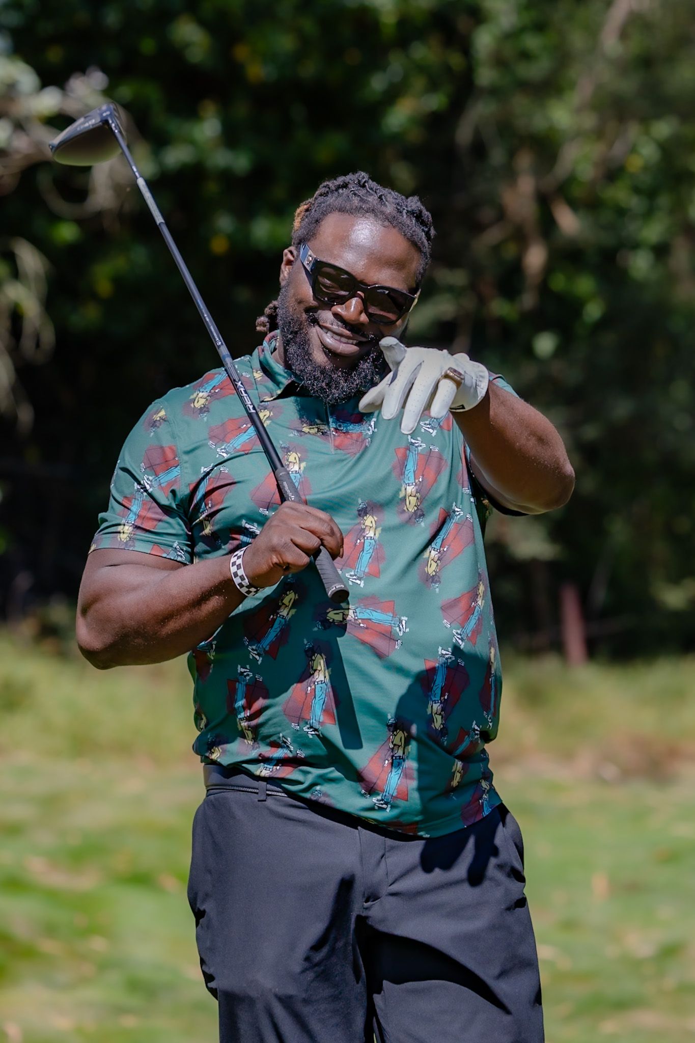 Smiling Black man in patterned golf shirt and sunglasses holding a golf club on a sunny golf course.