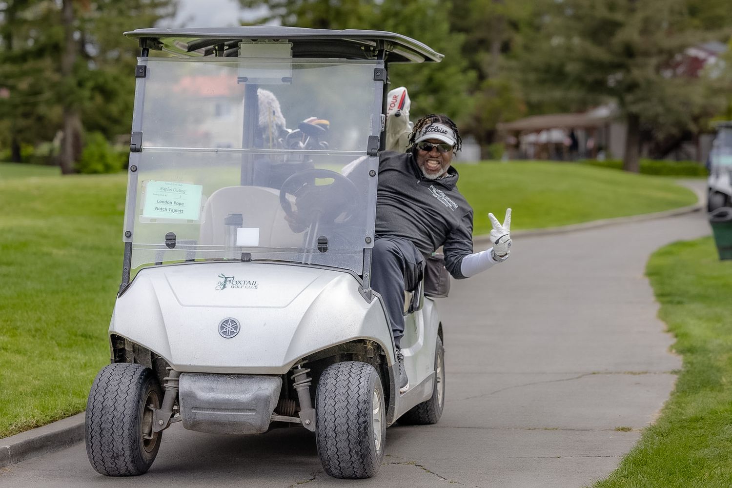 A cheerful golfer flashing a peace sign while riding a white Yamaha golf cart on a paved path at a golf course.